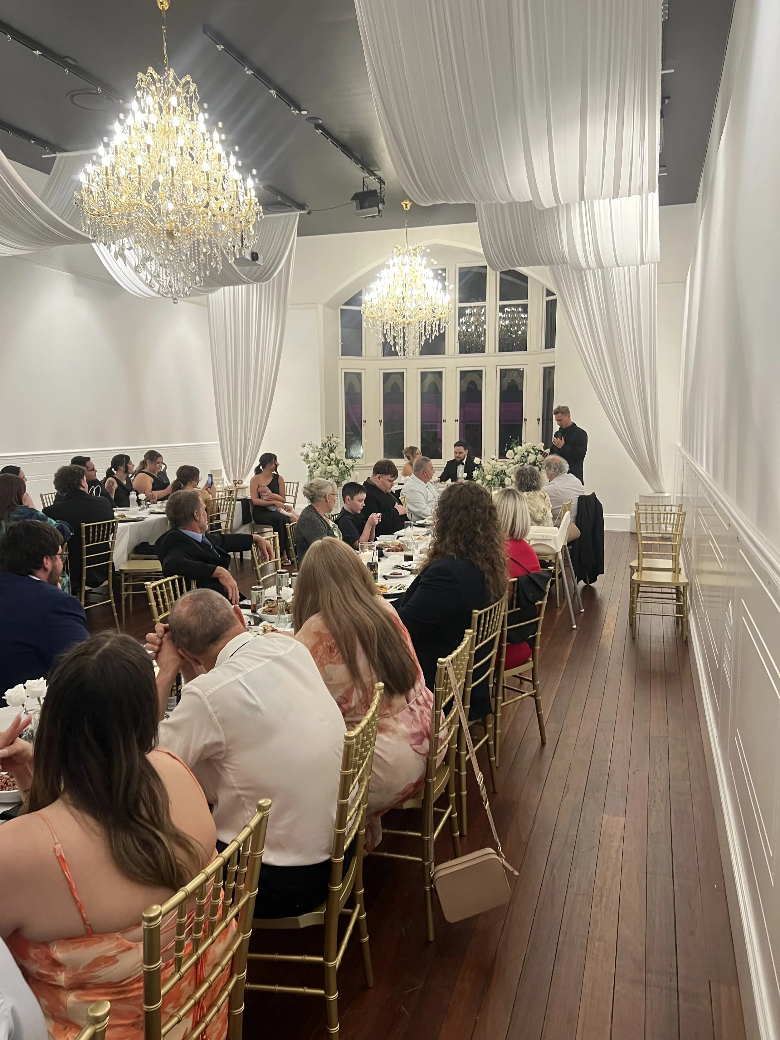 People seated at a long banquet table during a formal event in an elegant white room with chandeliers and large windows.