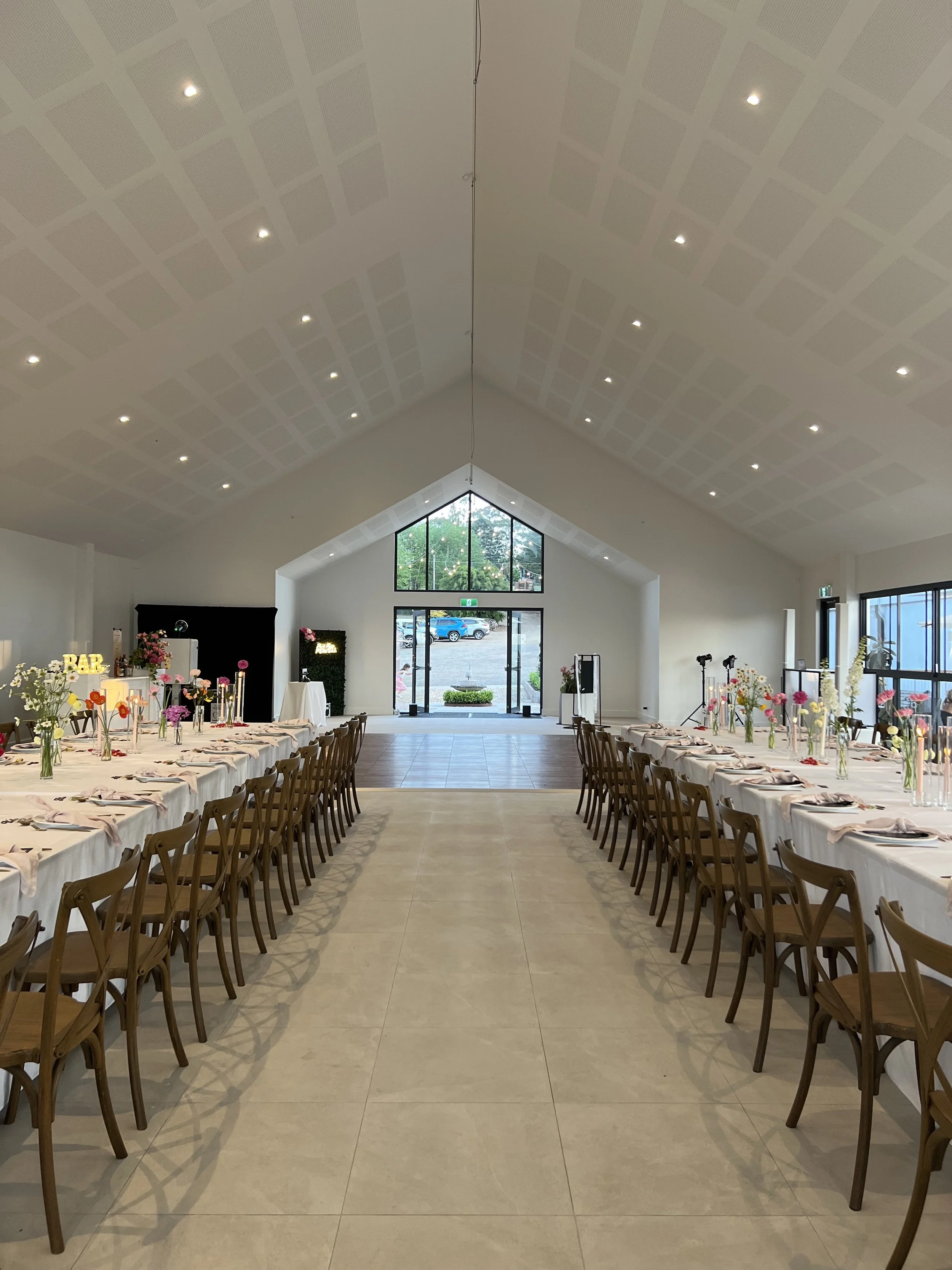 Event hall with two long tables set for a meal, decorated with pink and white flowers in vases, candles, and table settings. The hall has a high ceiling with recessed lighting, large windows, and a glass door at the front showing an outdoor parking lot and trees.