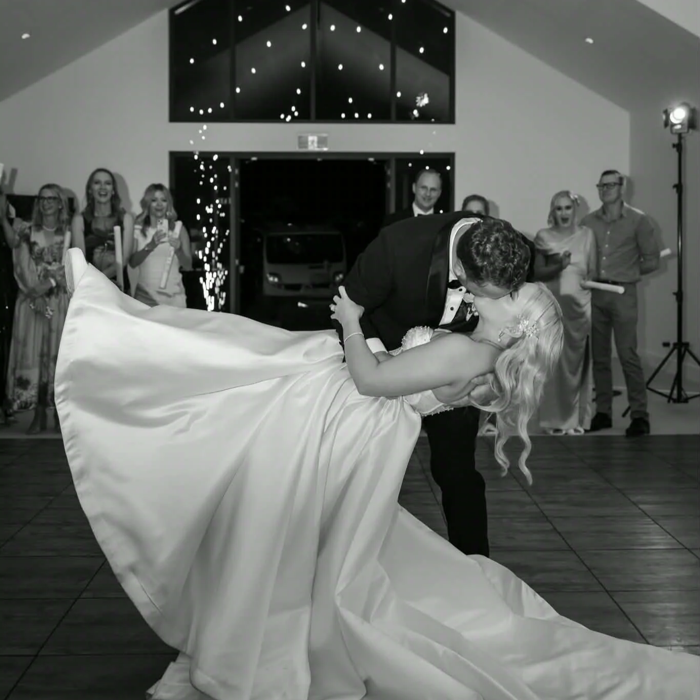 A couple shares a kiss during their wedding reception while the groom dips the bride, who wears a flowing wedding gown, in front of smiling guests.
