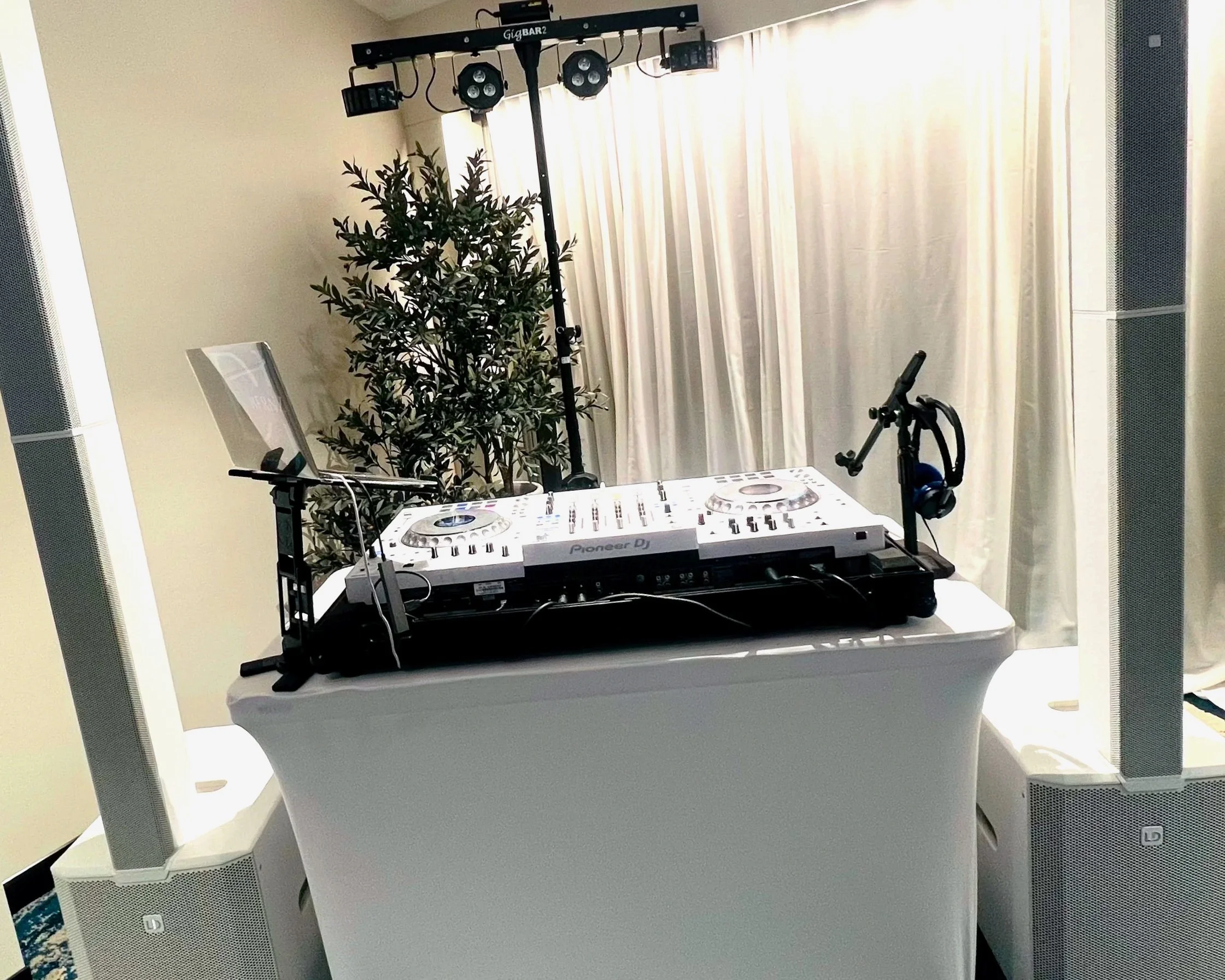 DJ setup with Pioneer DJ controller, laptop, headphones, and speakers in a room with curtains and a potted plant.