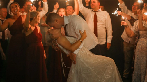 Couple sharing a kiss at a celebration with friends, some holding sparklers.