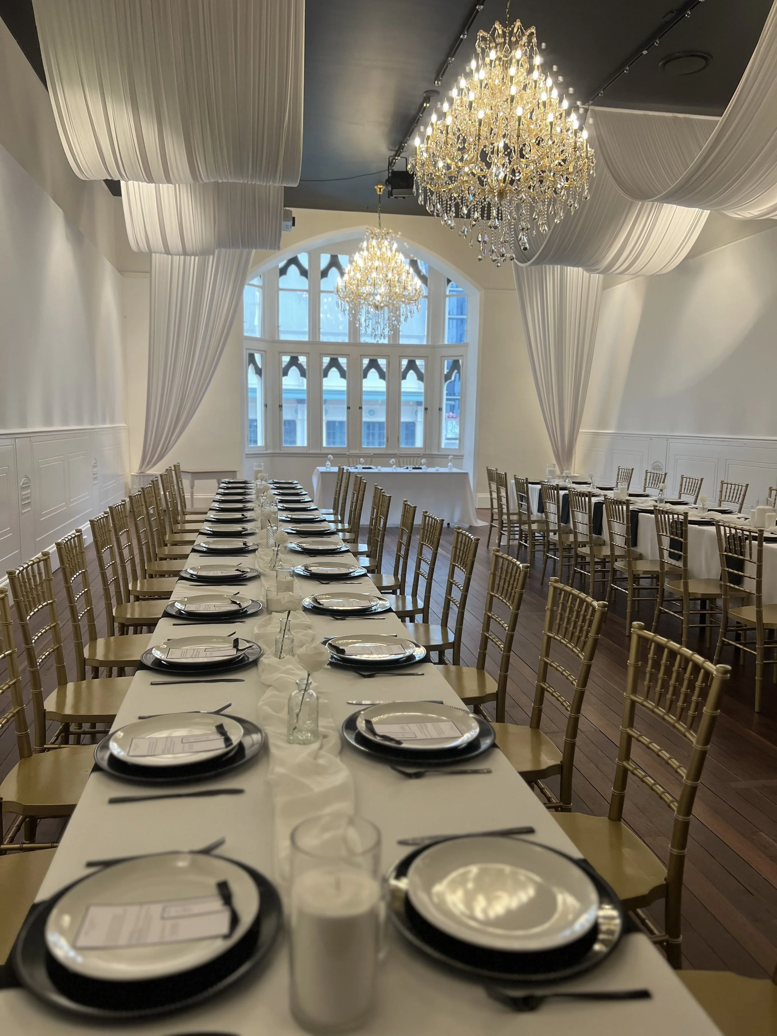 Elegant banquet hall with long table set for a formal event, decorated with white tablecloths, black and white plates, glasses, black napkins, and gold chairs. Large chandeliers and draped white curtains adorn the ceiling; a large window at the end of the room offers natural light.