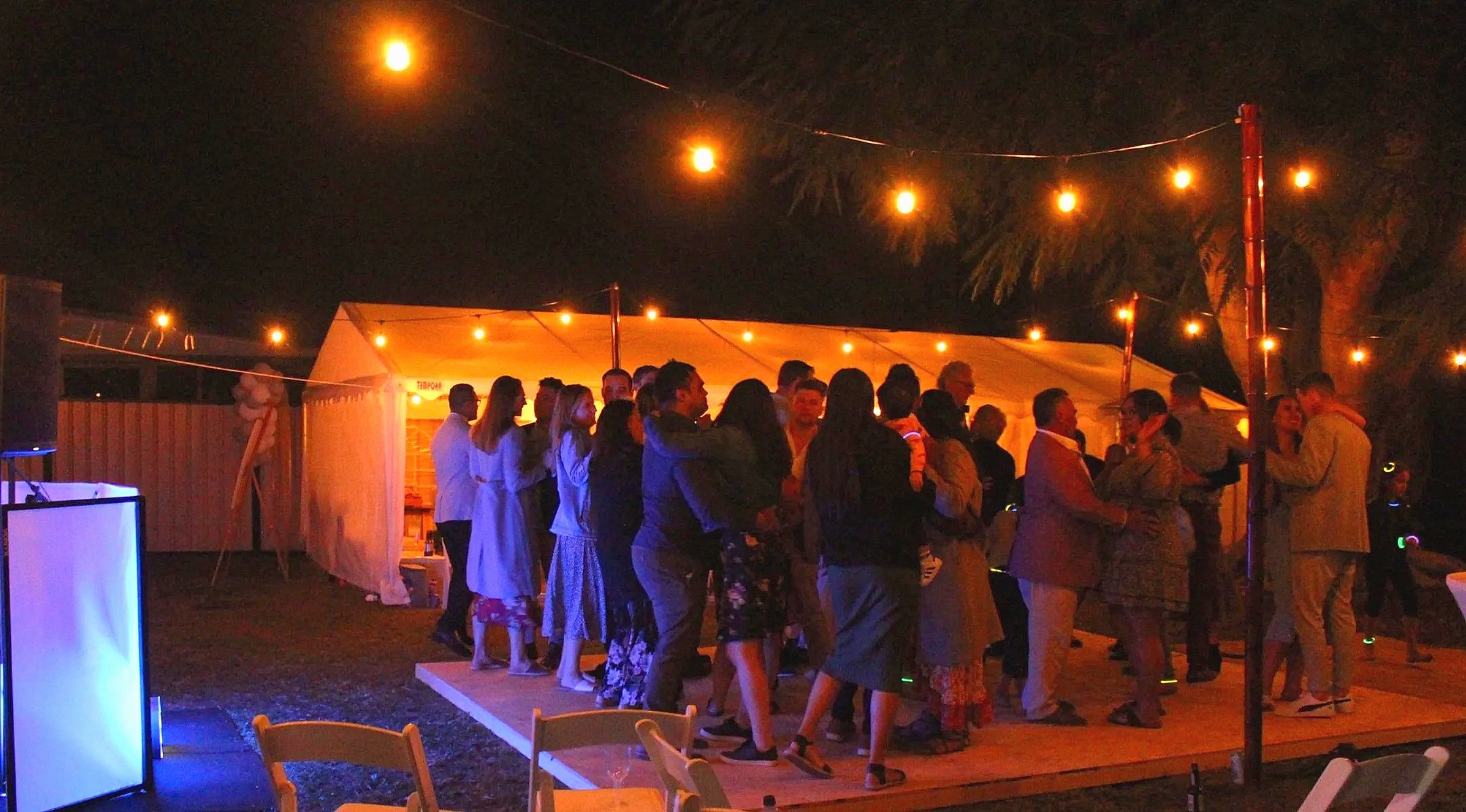 People dancing and socializing on a wooden dance floor outside at night, illuminated by string lights, with a tent in the background and trees visible.
