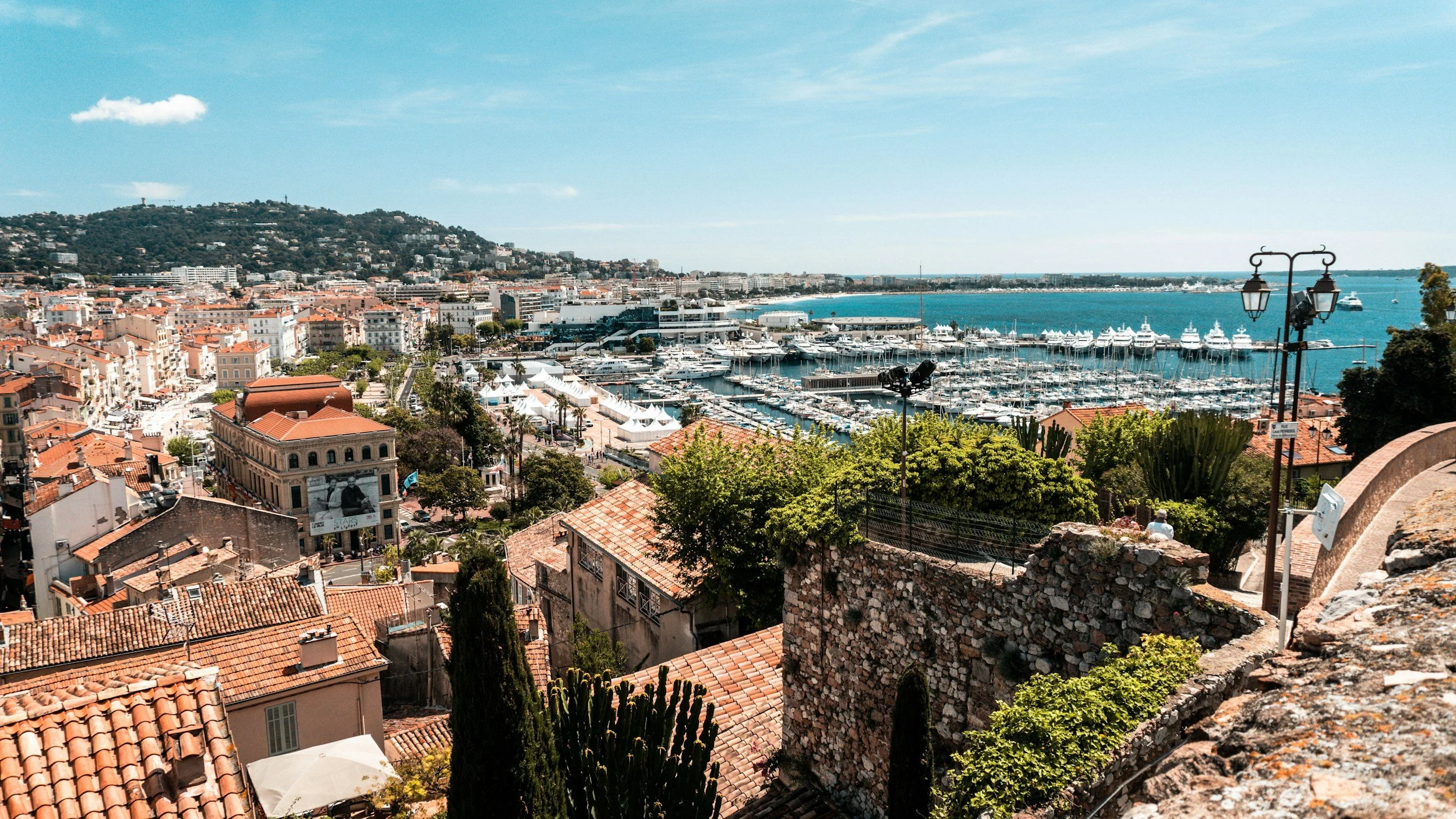 Aerial view of a European coastal city with historic waterfront architecture, boats anchored in turquoise water, and a scenic marina, representing international film festival destinations and global travel for filmmakers.