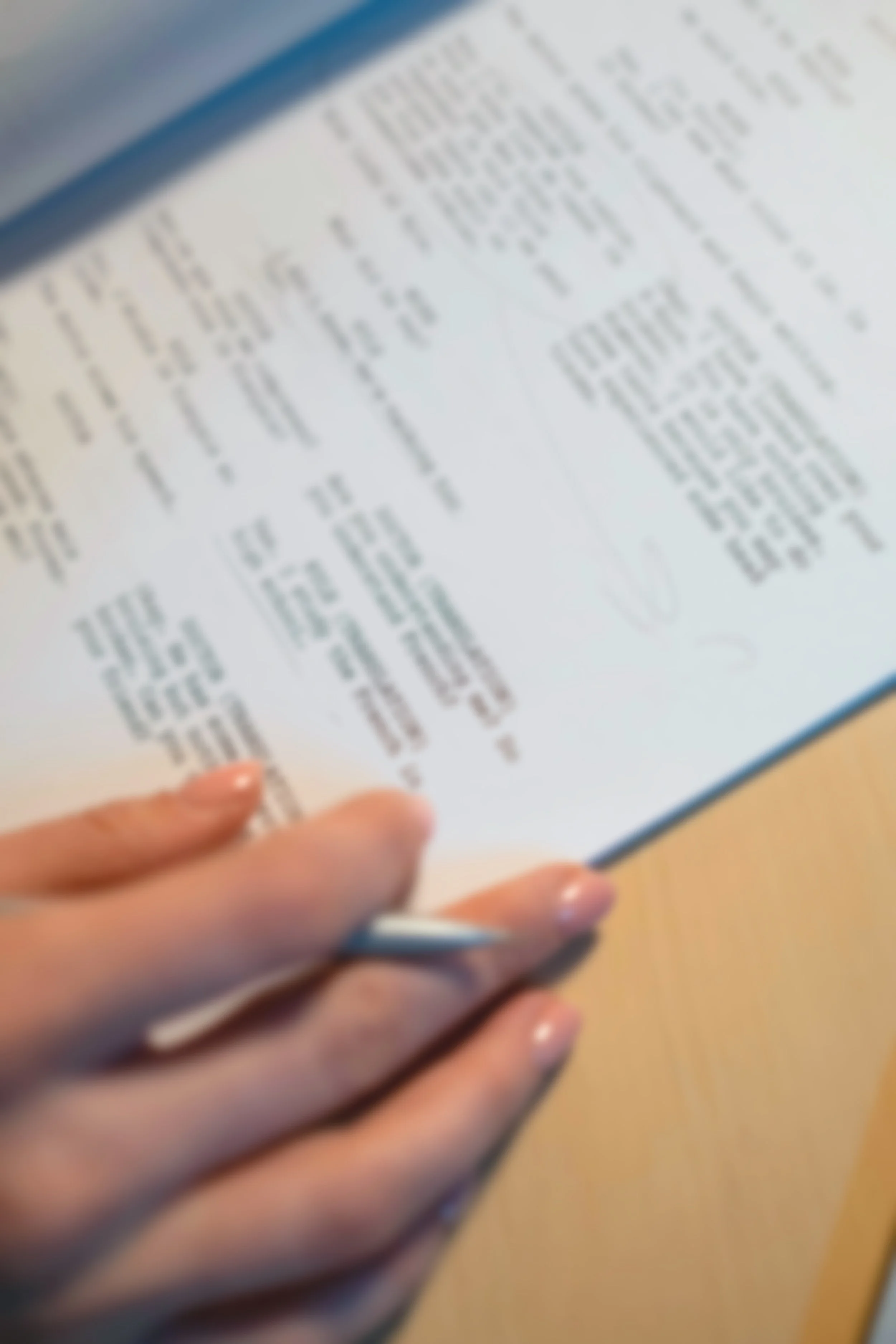 A person's hand holding a pen over a blurred page of text, possibly in a book or document, on a wooden surface.