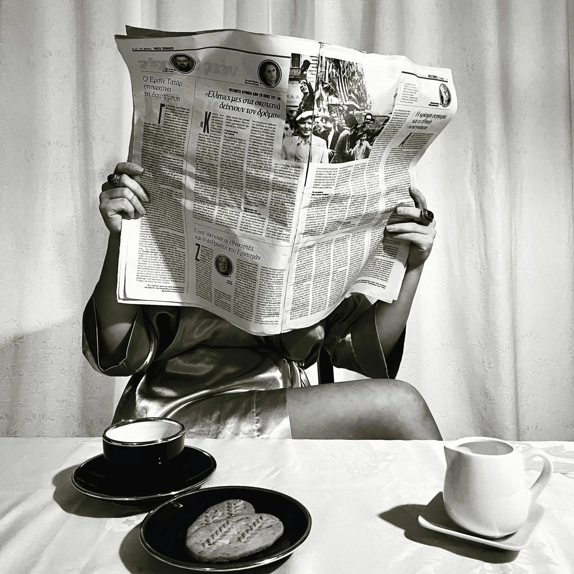 Monochrome image of a person holding a newspaper at a breakfast table with coffee and food, representing ordinary domestic life used as a backdrop in modern film and TV narratives
