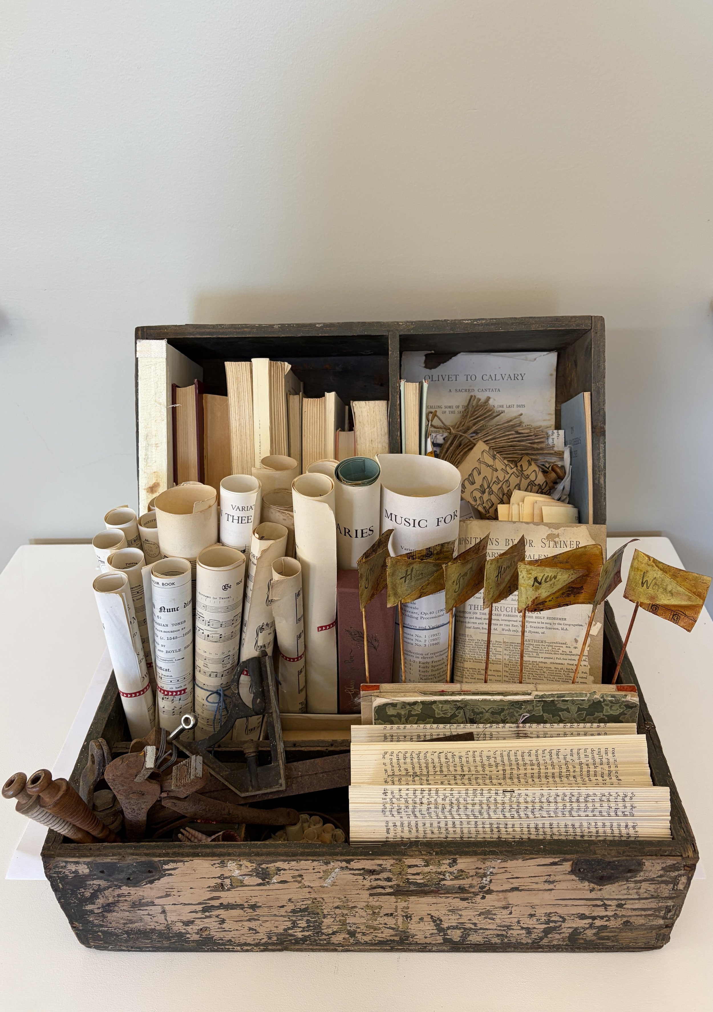 Booksmith’s Toolbox gathers the discarded tools and materials of bookmaking into a cabinet of curiosities. Old books, fragments of music, rolled pages, rusted tools, thread and handmade paper are assembled in a weathered carpenter’s box, suggesting a