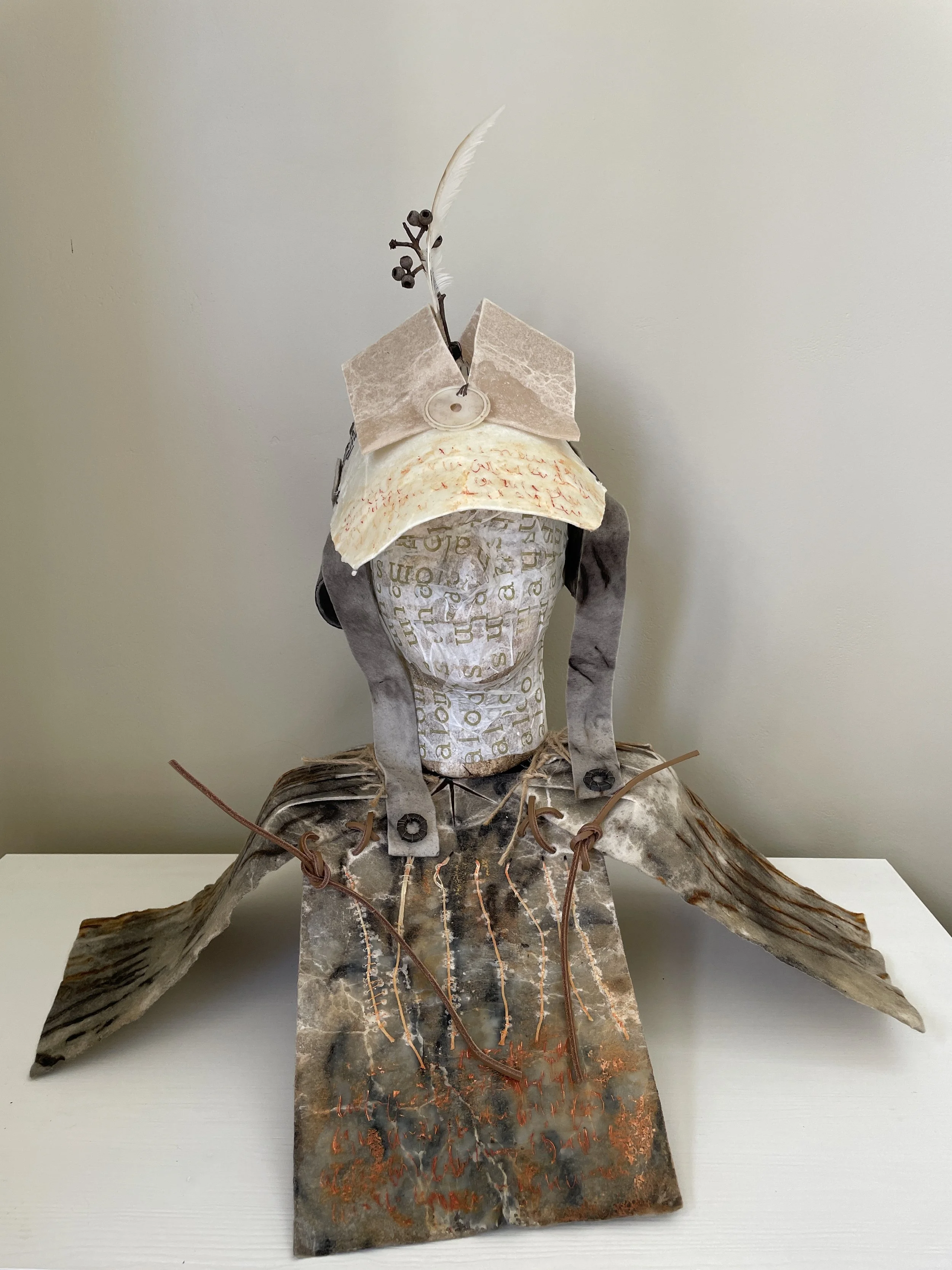 The figure-like presence stands as a vessel of history and human gesture. The layered papers and stitched seams evoke garments or armour, carrying the markings of spoken prayers layered over time. The feather and botanical element rising from the top