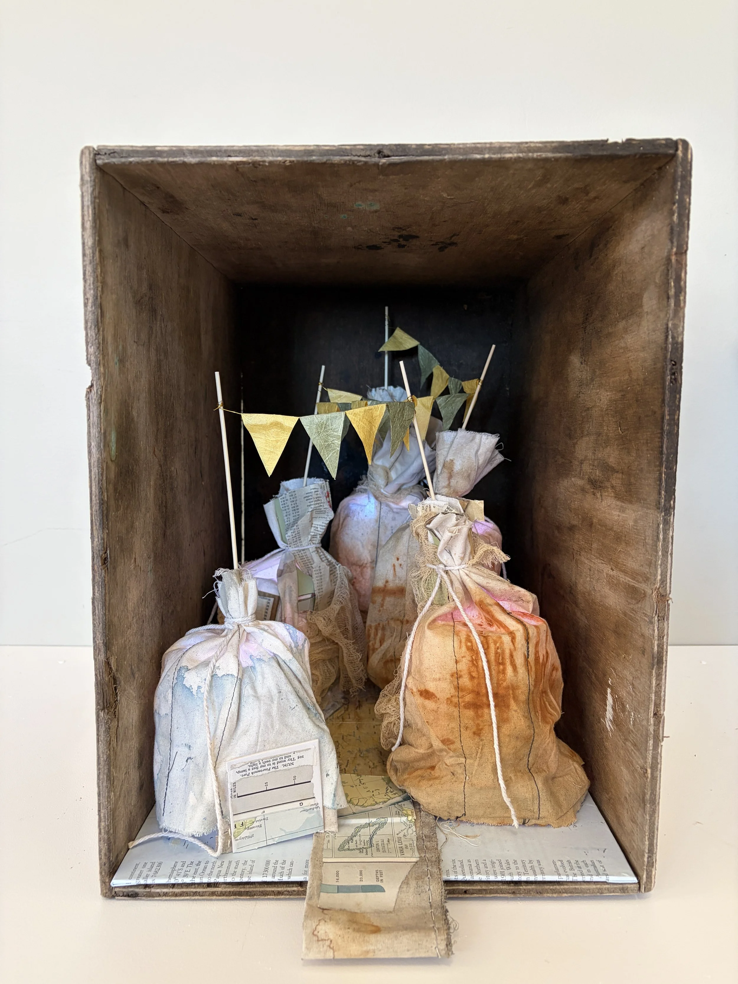 To my feet a lamp takes its title from Psalm 119 and its meditation on God’s word as a guide for the journey. Four cloth-covered lamps stand within an old wooden box, connected by a string of small flags. Beneath them, fragments of a world atlas sugg