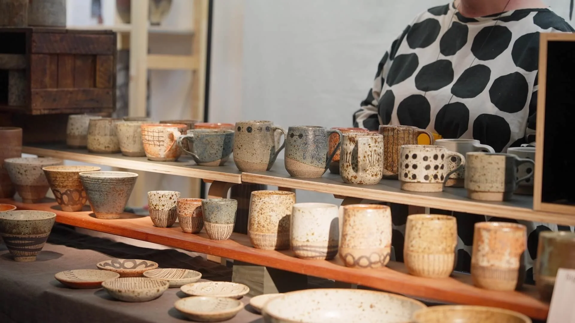 Display of handmade ceramic cups, bowls, and plates on a multi-tiered wooden table at a craft fair.