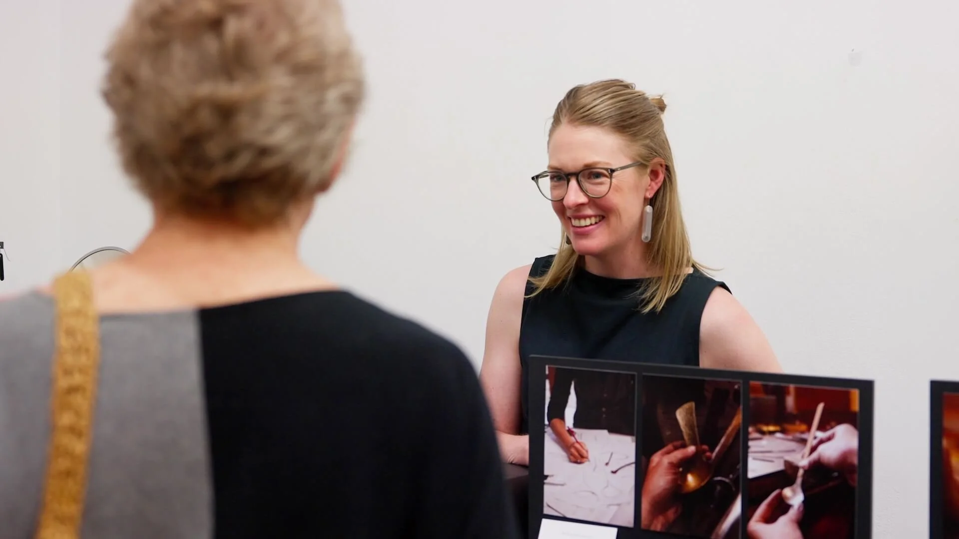 Two women having a conversation, one with glasses and earrings, smiling, at an exhibition table displaying photos of hands holding various objects.
