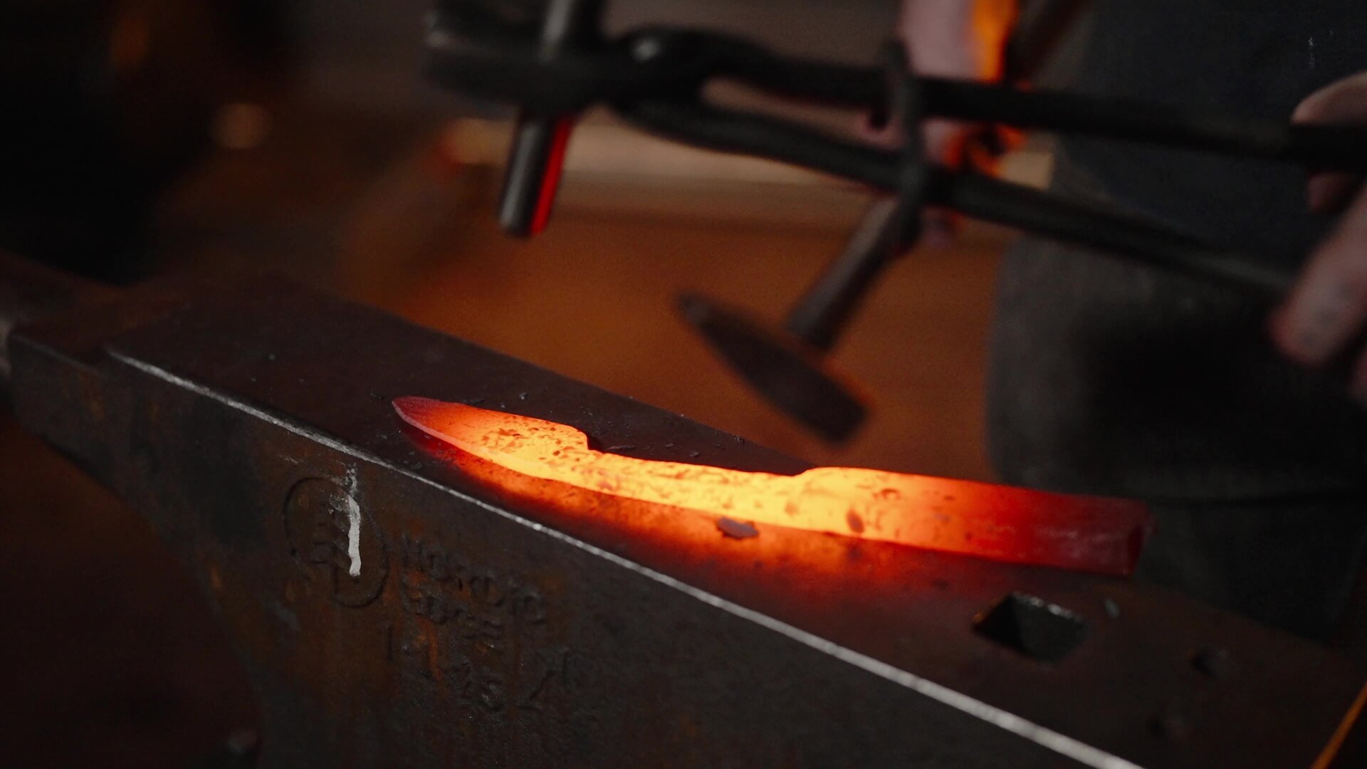 A glowing piece of hot metal is being worked on a blacksmith anvil, with a hammer nearby. The area is dimly lit, showing the bright orange and red heat of the metal.