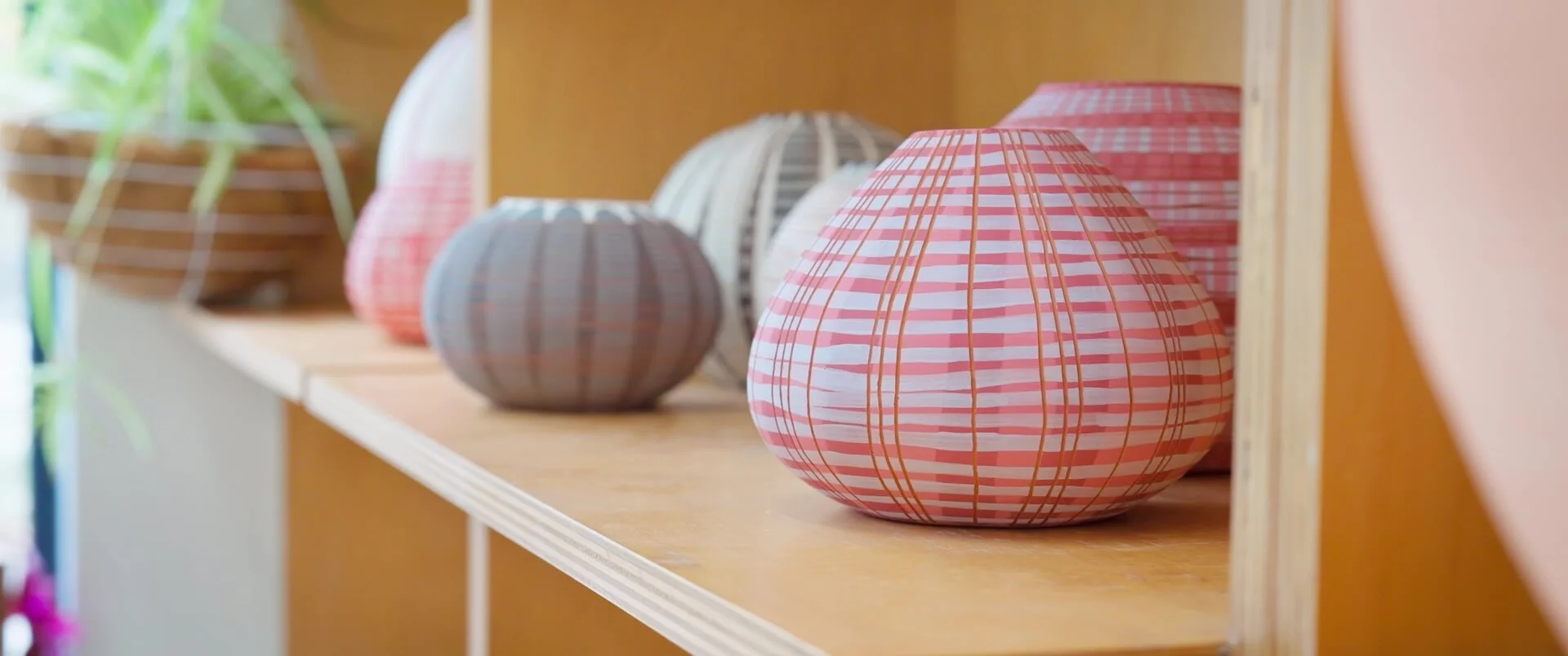 Colorful decorative paper lanterns in pink, gray, and white on a wooden shelf in a bright room.