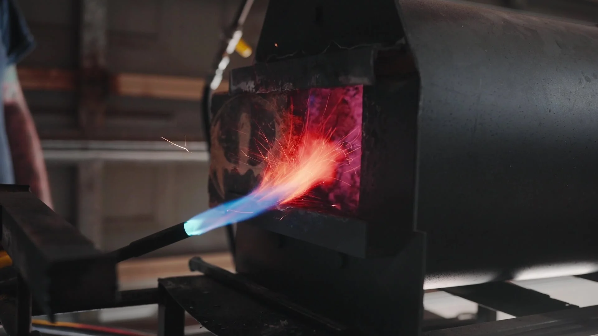 A close-up of a blowtorch igniting a gas-forge, with a bright orange and blue flame,.