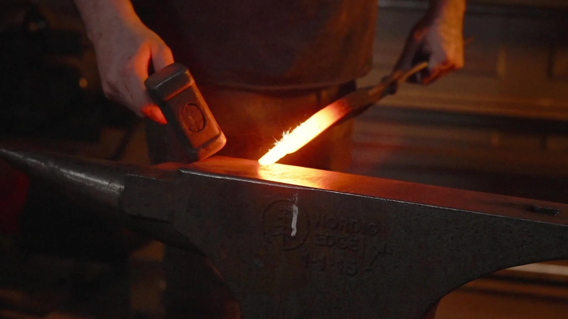 A blacksmith, Dan Venables, inspects a glowing hot knife on his anvil, hammer at the ready to make the next strikes to shape it.