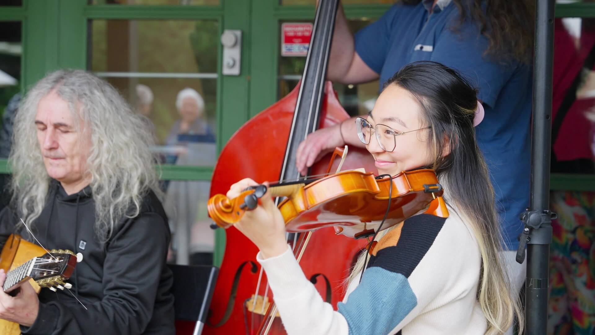 Young woman playing the violin in a group, with a man playing the guitar beside her and a bassist in the background.