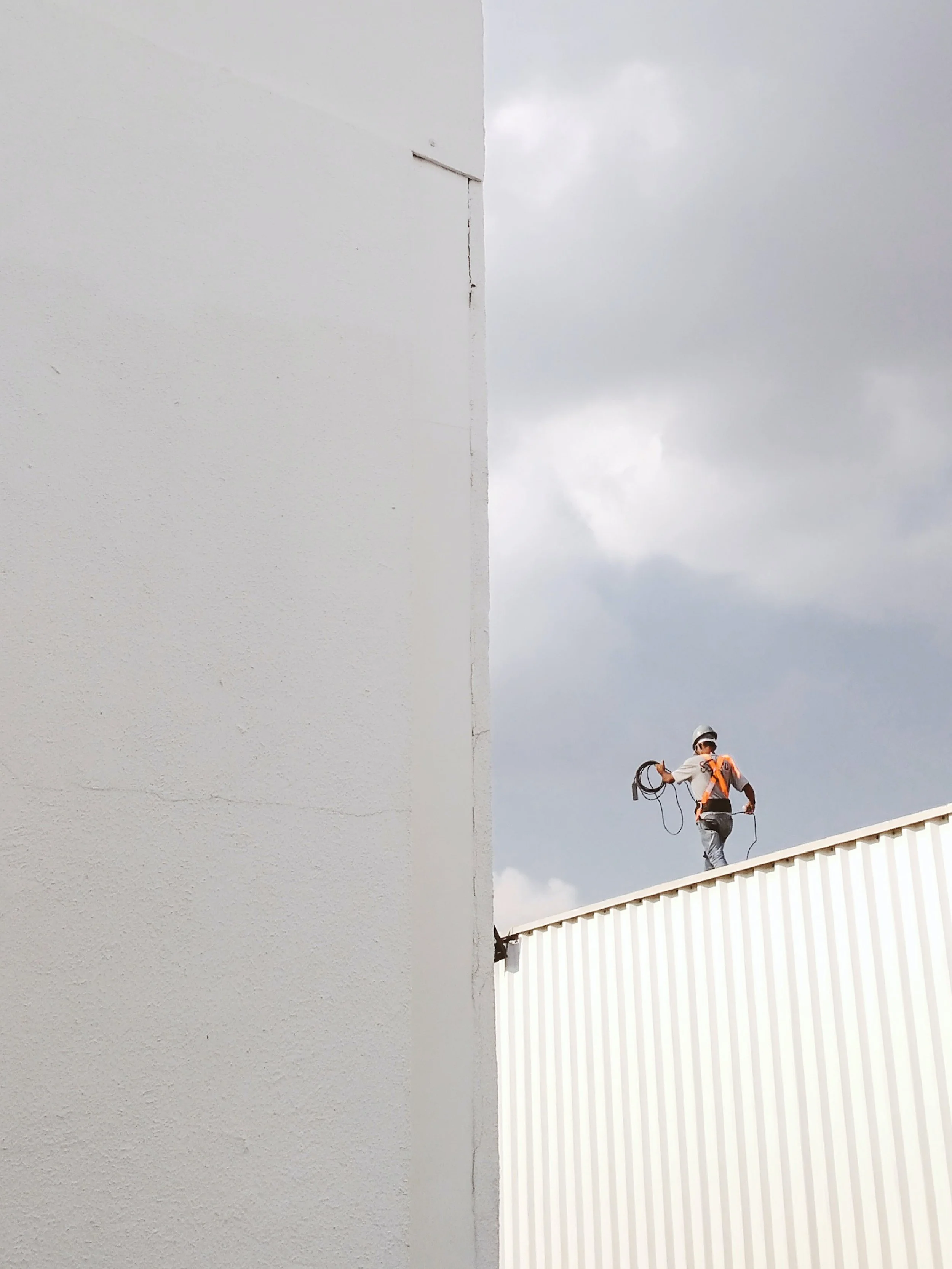 A worker in safety gear walking on a ledge outside a building, with a large white wall on the left and a gray cloudy sky in the background.
