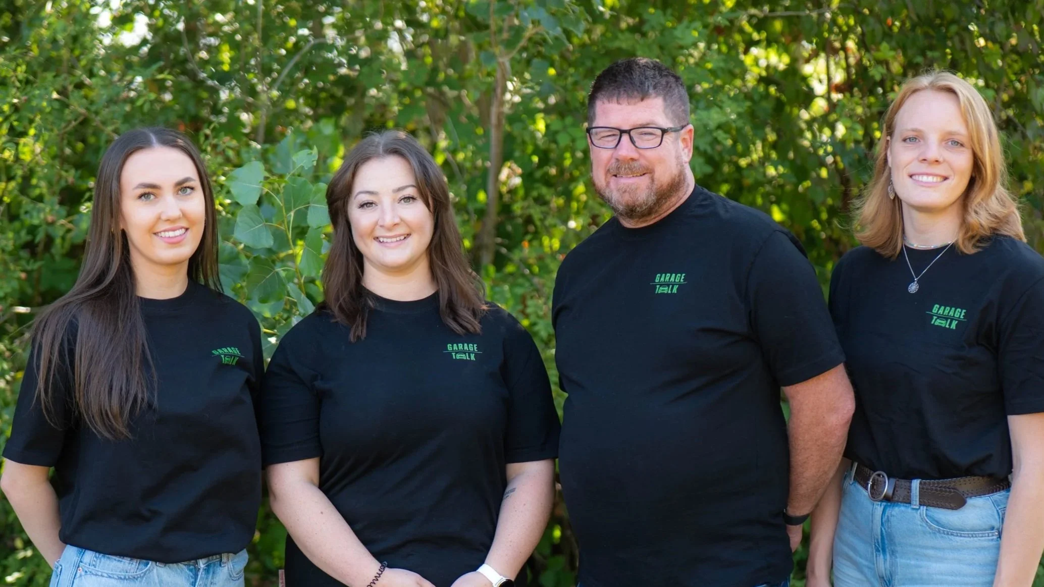 Group of four people standing outdoors with green trees in the background, wearing black shirts with green text that says 'Garage Talk'.