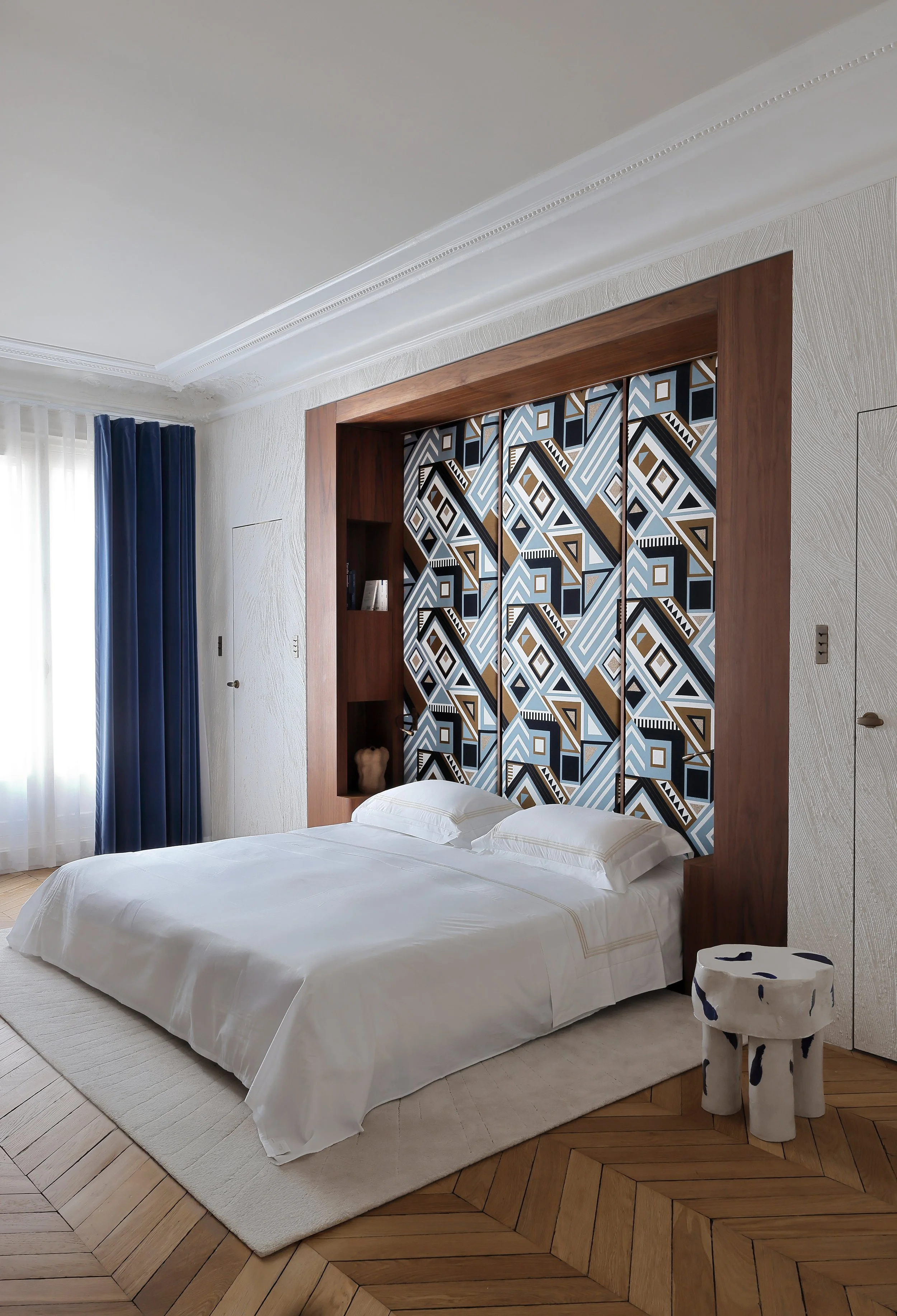 Chambre avec lit blanc, tête de lit en bois avec motif géométrique moderne, sol en parquet, fenêtre avec rideaux bleus et blancs, petit banc blanc avec taches bleues.