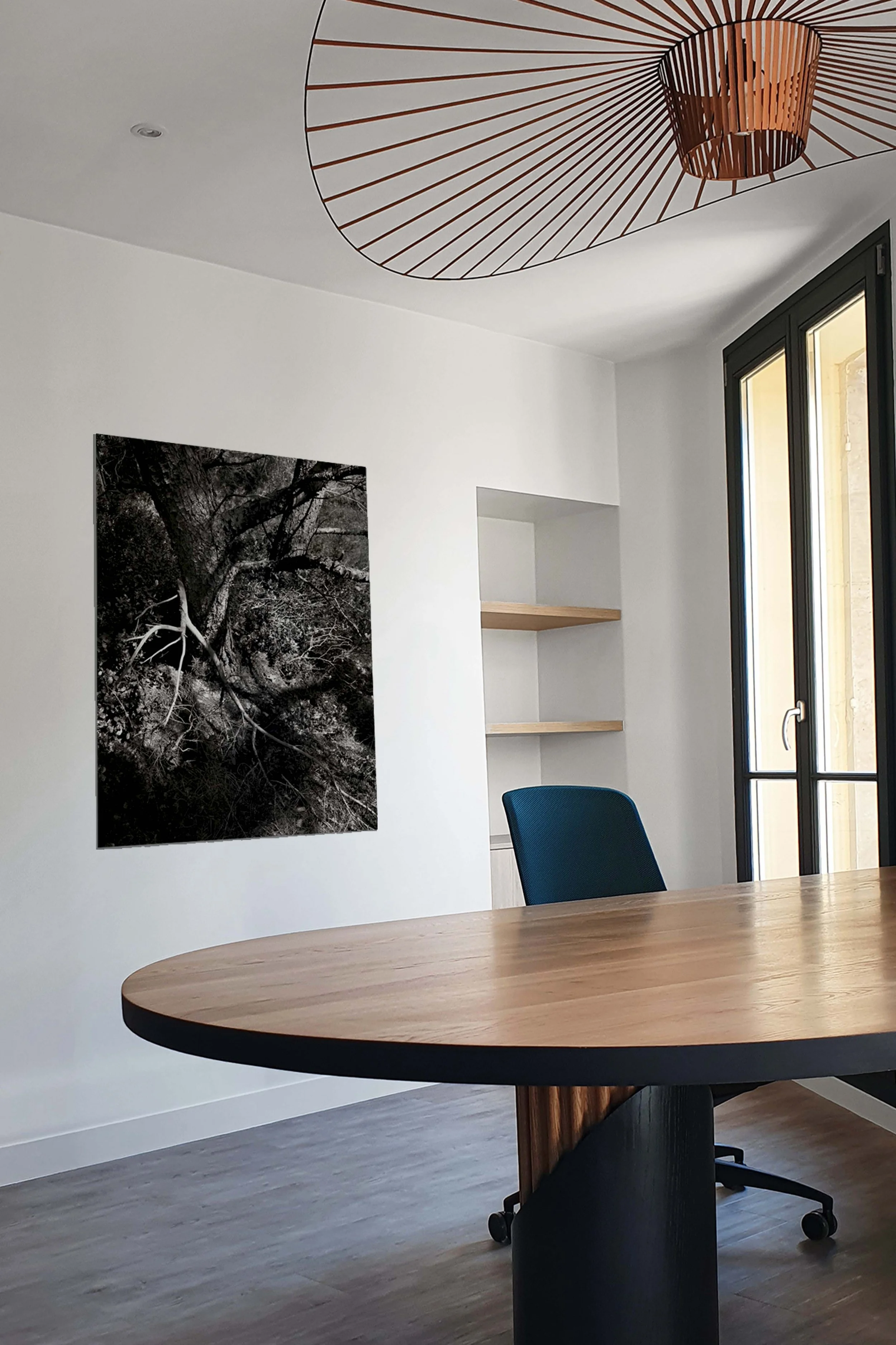 Meeting room or office with round wooden table, blue chair, black-and-white portrait of a tree, wooden shelves on the wall, double-sash window letting in the light.