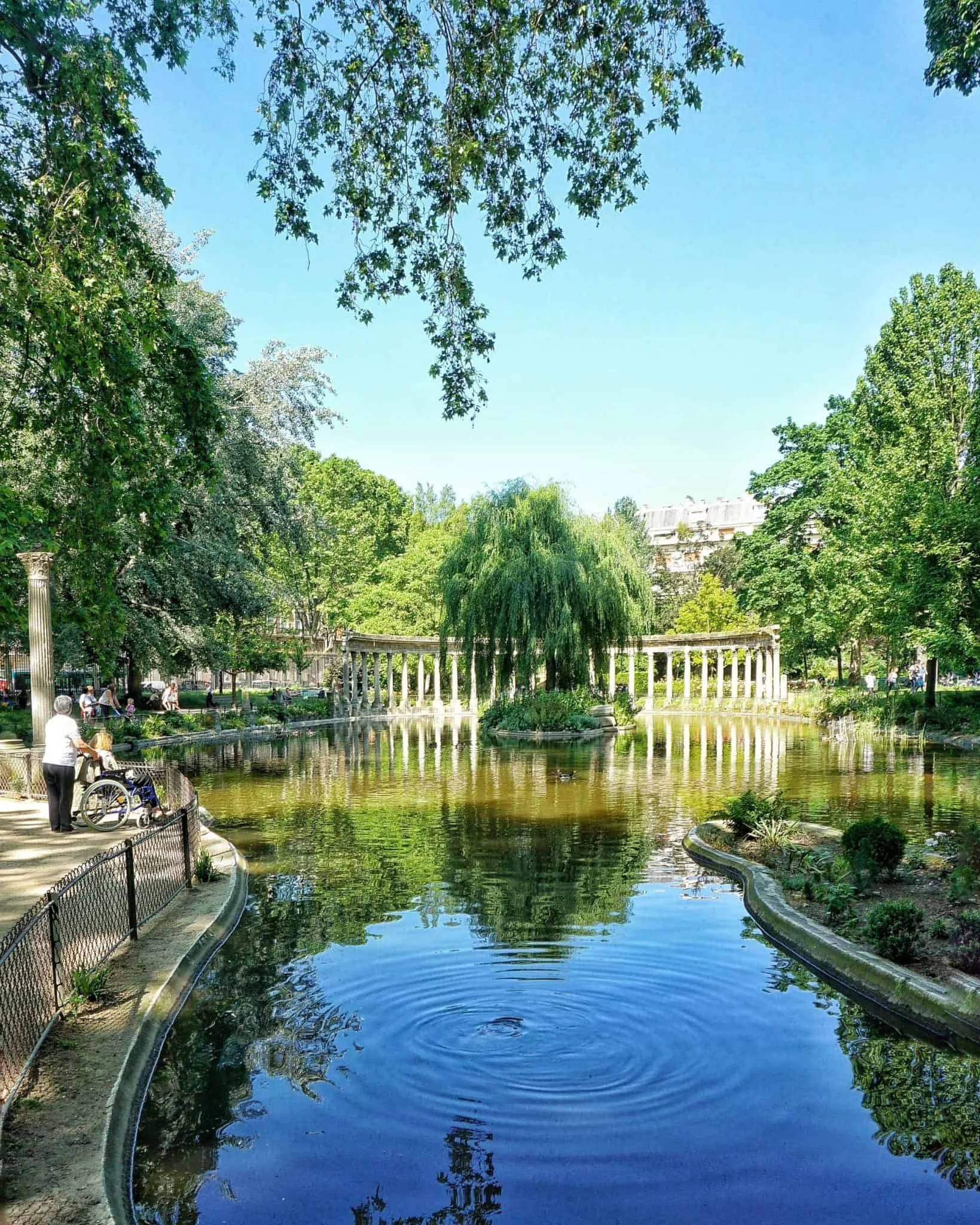 Parc avec un étang, arbres verts, un chemin bordé d'une barrière, personnes se promenant, colonnes blanches en arrière-plan, ciel clair, ambiance paisible.