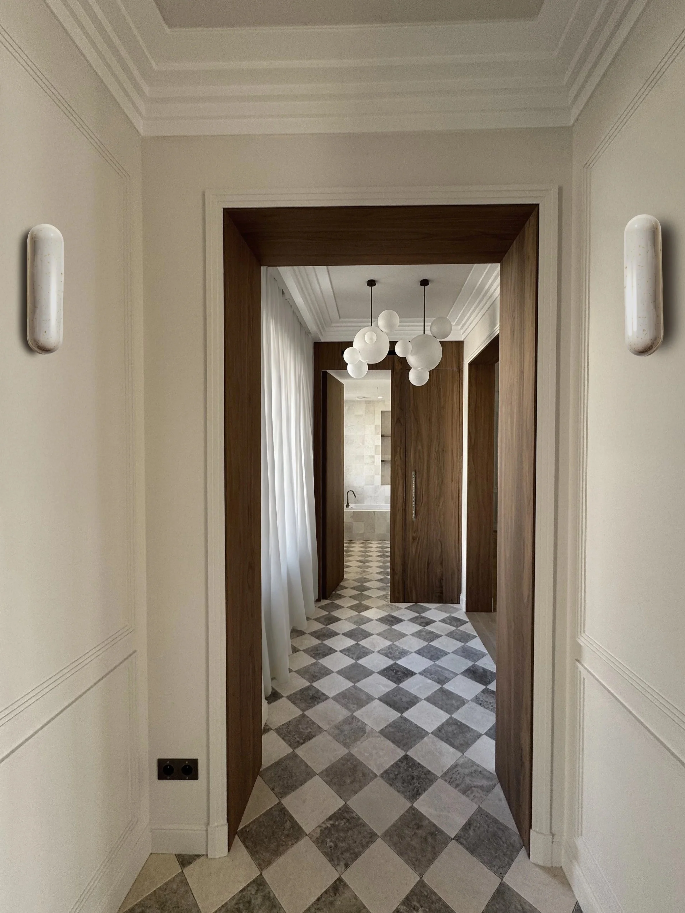 View of an interior hallway with gray and white tiled floor, light-colored walls, walls with moldings, wooden doors, pendant light fixtures with white globes.