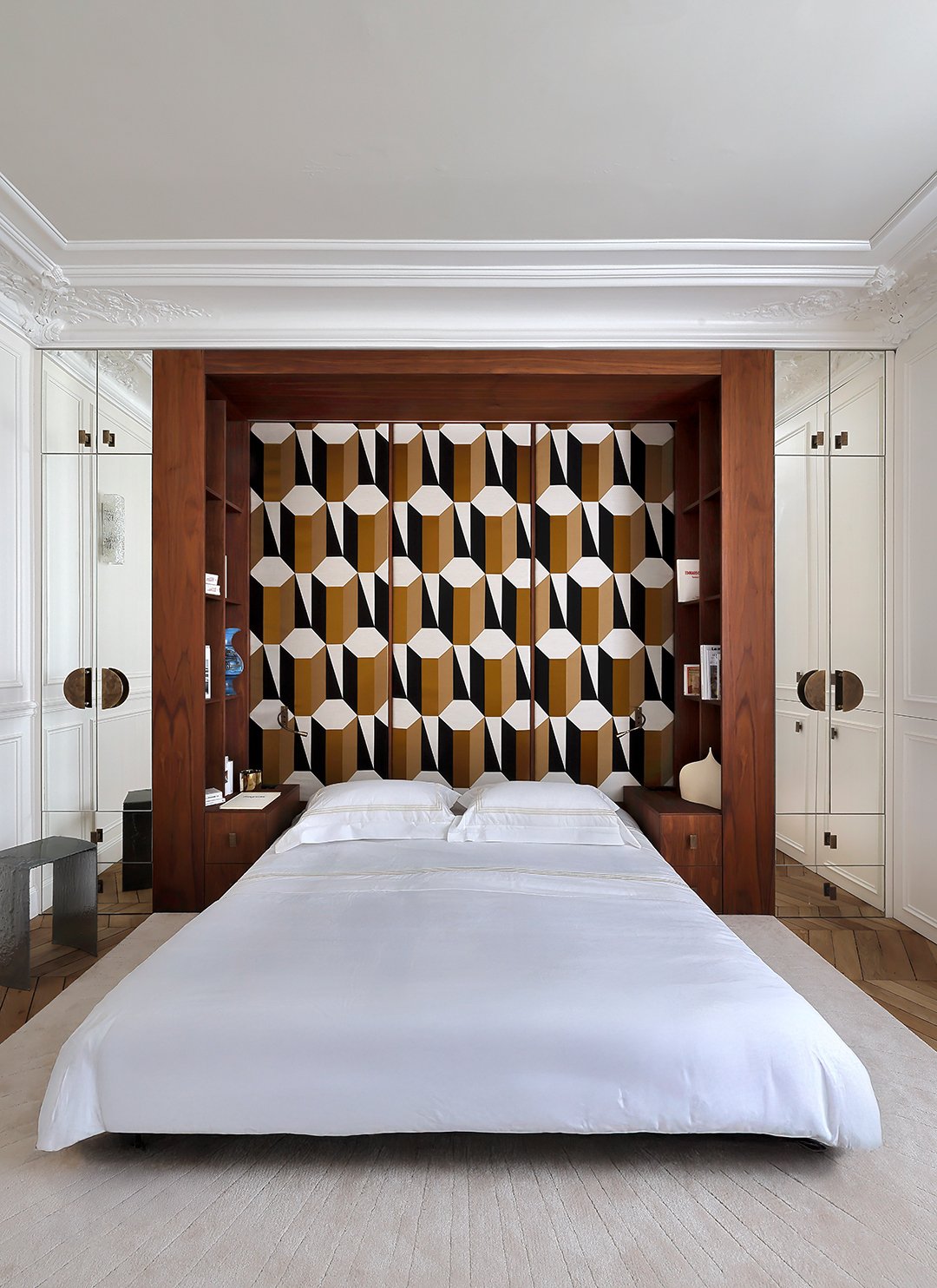 Chambre moderne avec un lit blanc, un mur en papier peint avec un motif géométrique en noir, blanc et doré. Armoires blanches avec poignées en cercle, bois foncé encadrant le lit, et petite table de nuit en métal.