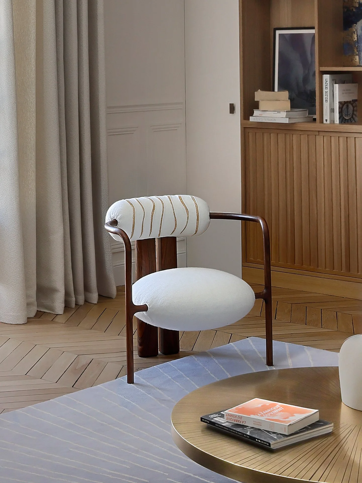 Une chaise élégante en bois avec un oreiller blanc à rayures or, placée dans un salon décoré avec un tapis, une table basse, des livres et une bibliothèque en bois.
