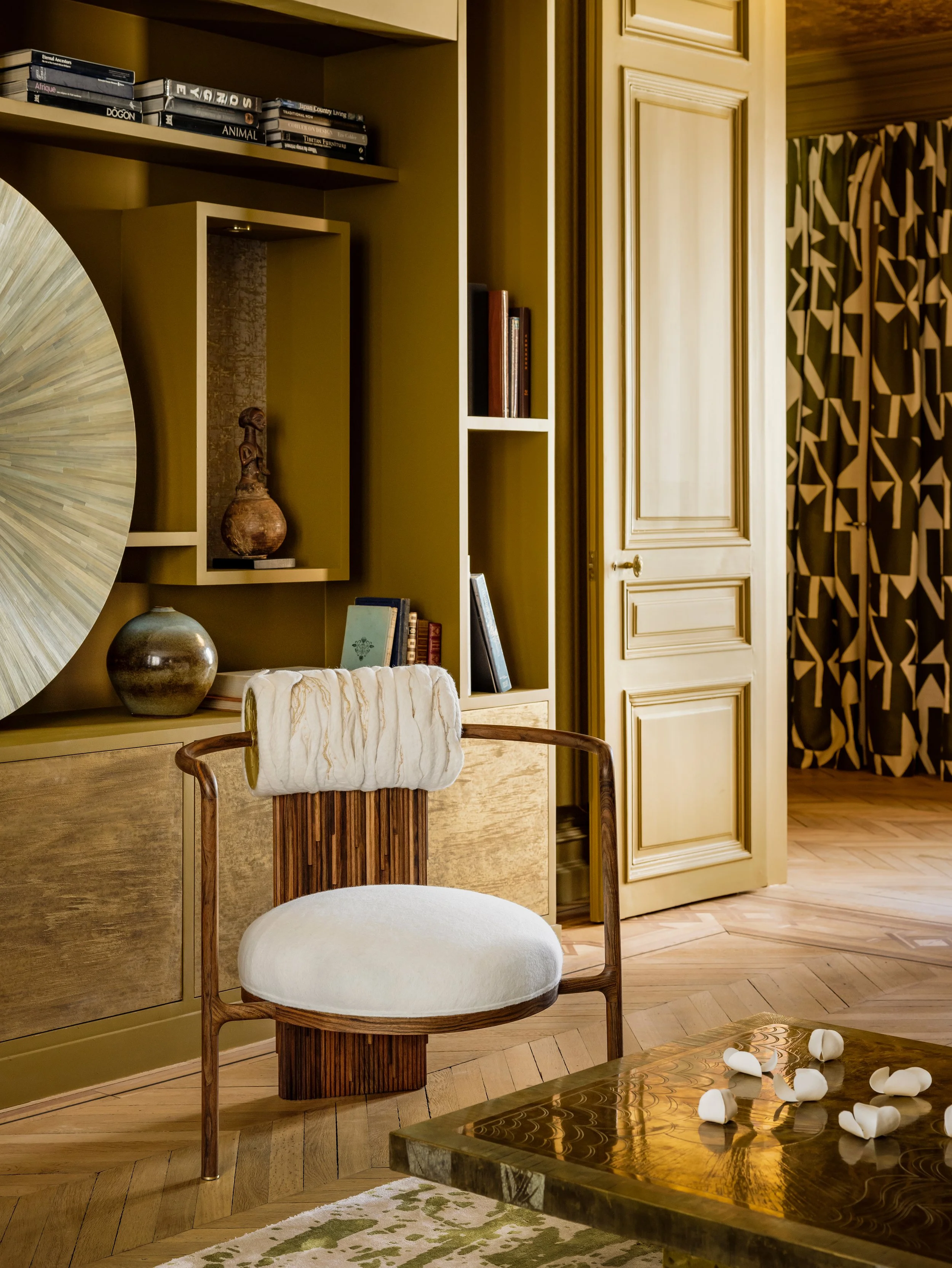 Intérieur d'un salon avec étagères contenant des livres, une porte ouverte, un fauteuil en bois avec assise et dossier en tissu clair, une table en bois brillant avec des rouleaux de papier toilette, et des rideaux avec un motif géométrique noir et beige.