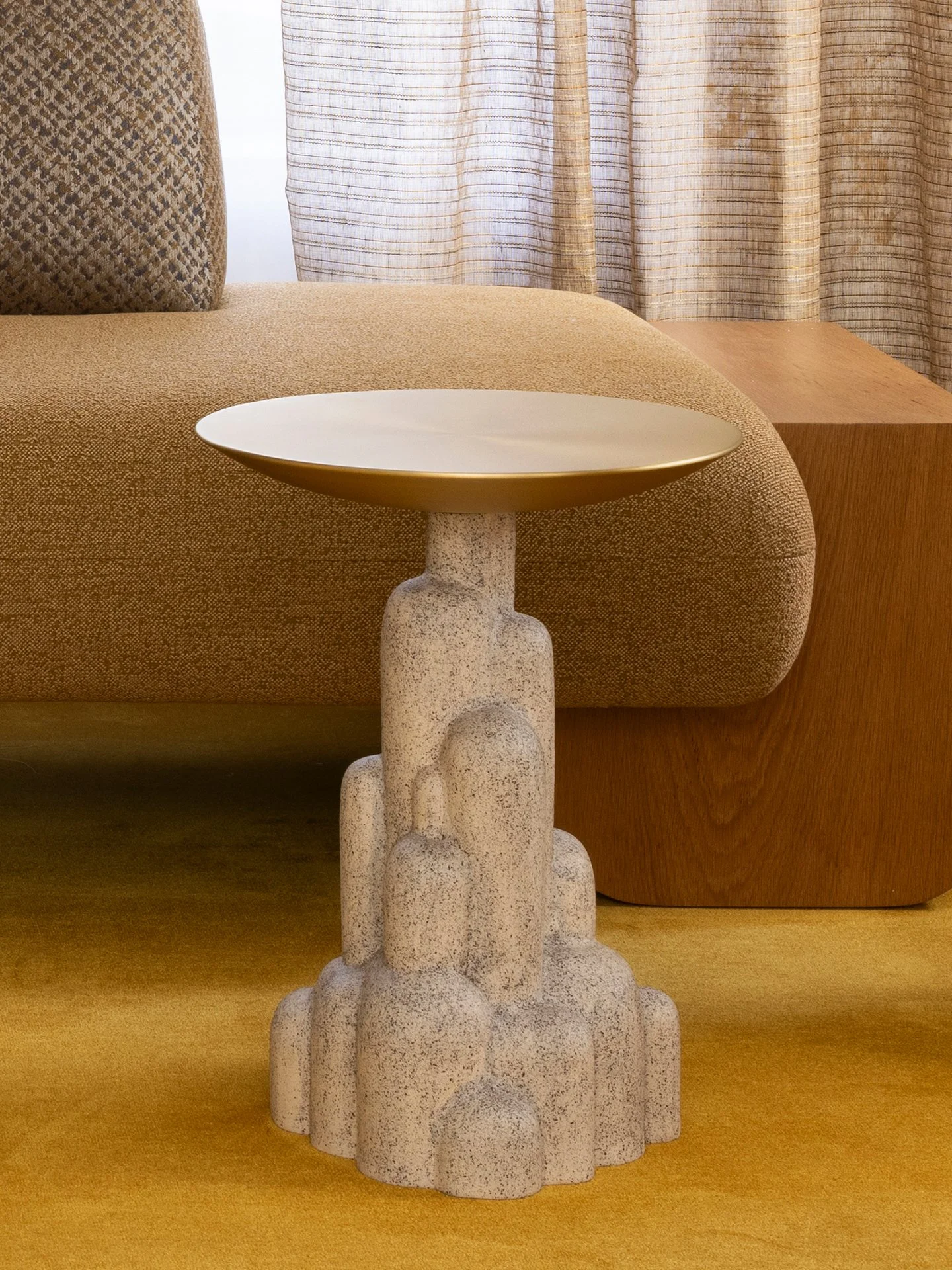 Une petite table avec un piètement en forme de figures abstraites en béton et un plateau en métal doré, placée devant un canapé beige et un rideau rayé dans une pièce moderne.