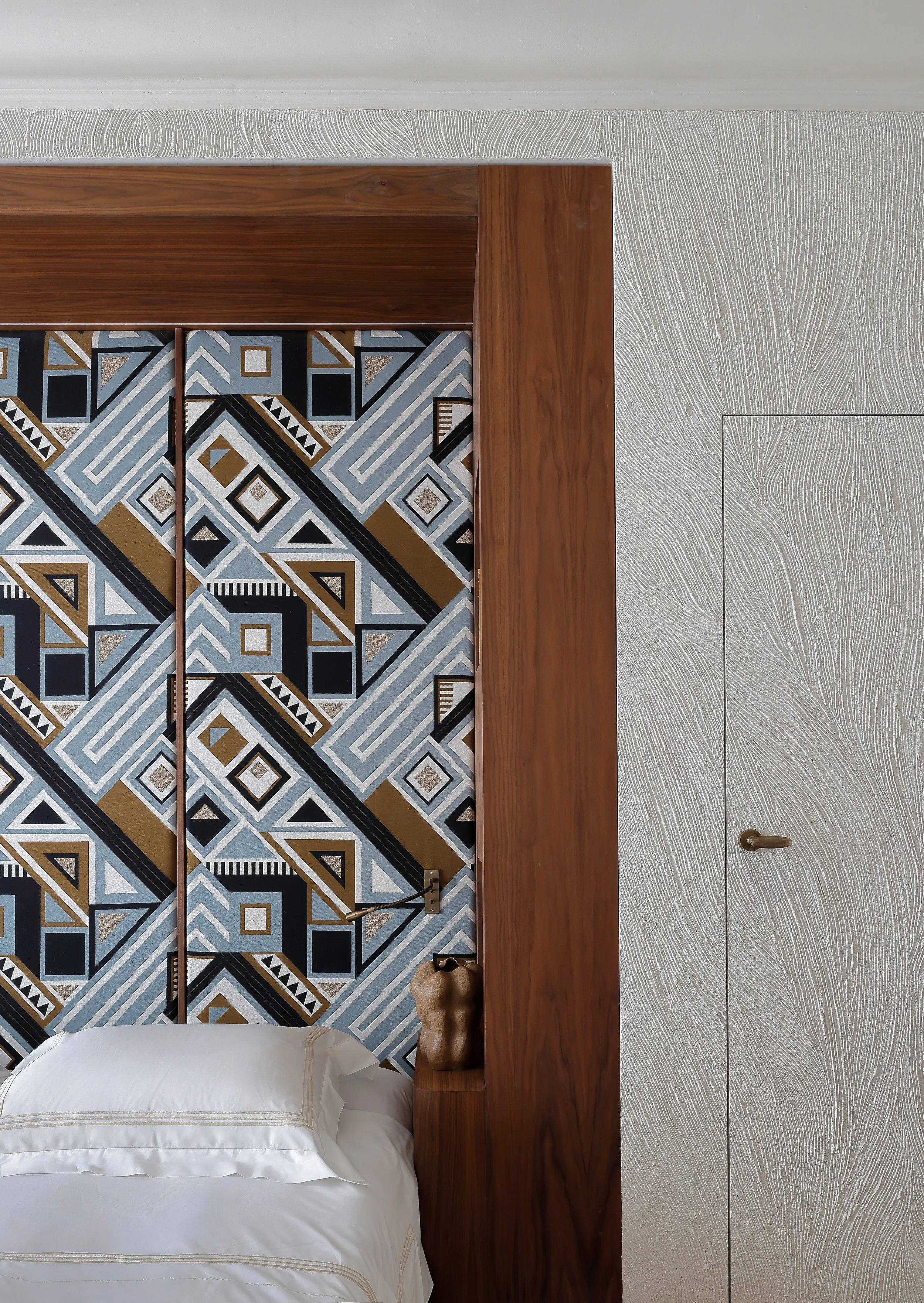Photo d'une chambre avec une tête de lit en bois, un coussin blanc, un motif géométrique sur le mur derrière la tête de lit et une porte à droite.
