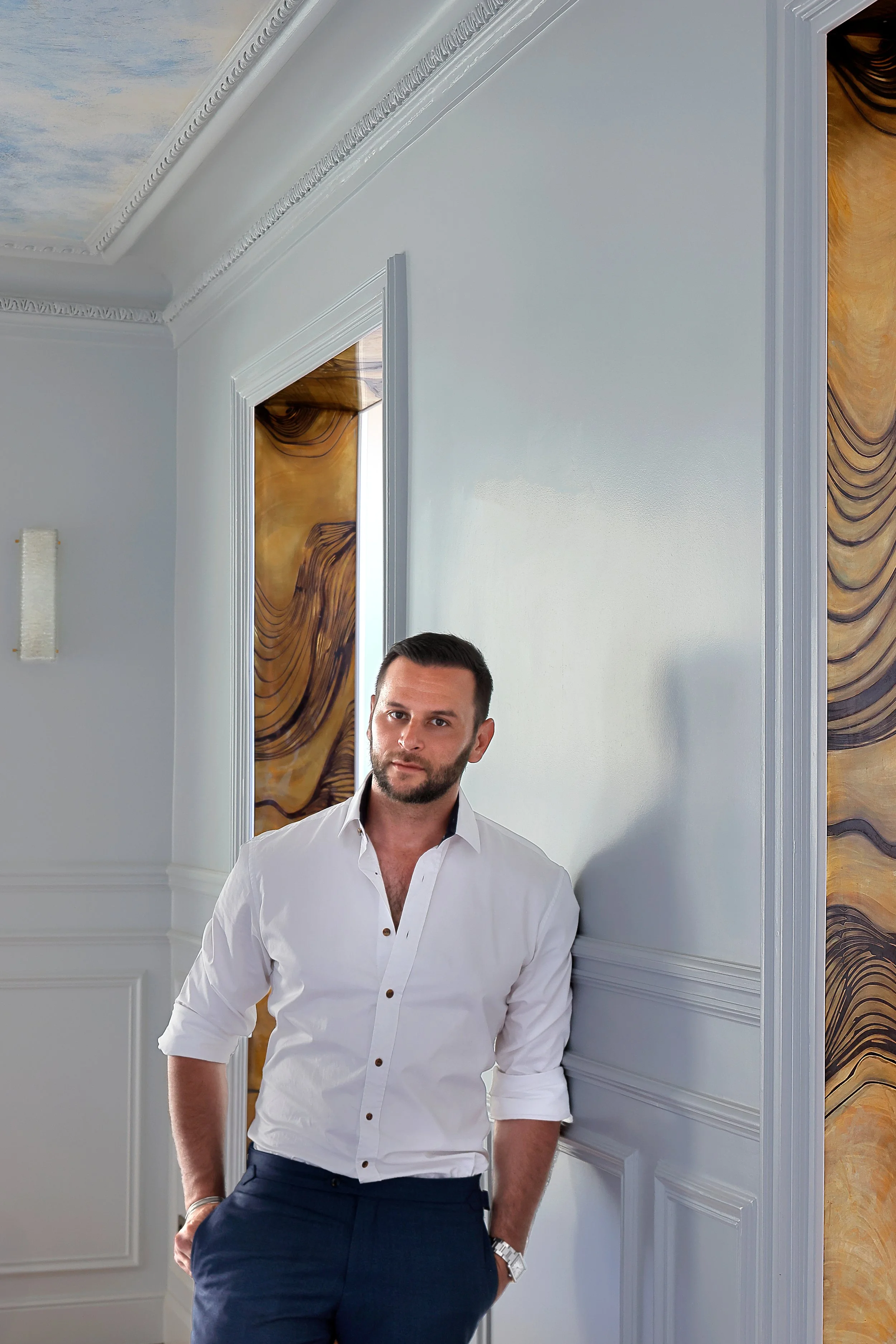 Homme en chemise blanche et pantalon bleu clair, debout contre un mur blanc dans une pièce élégante avec des détails en moulures et une décoration en marbre marron autour des portes.