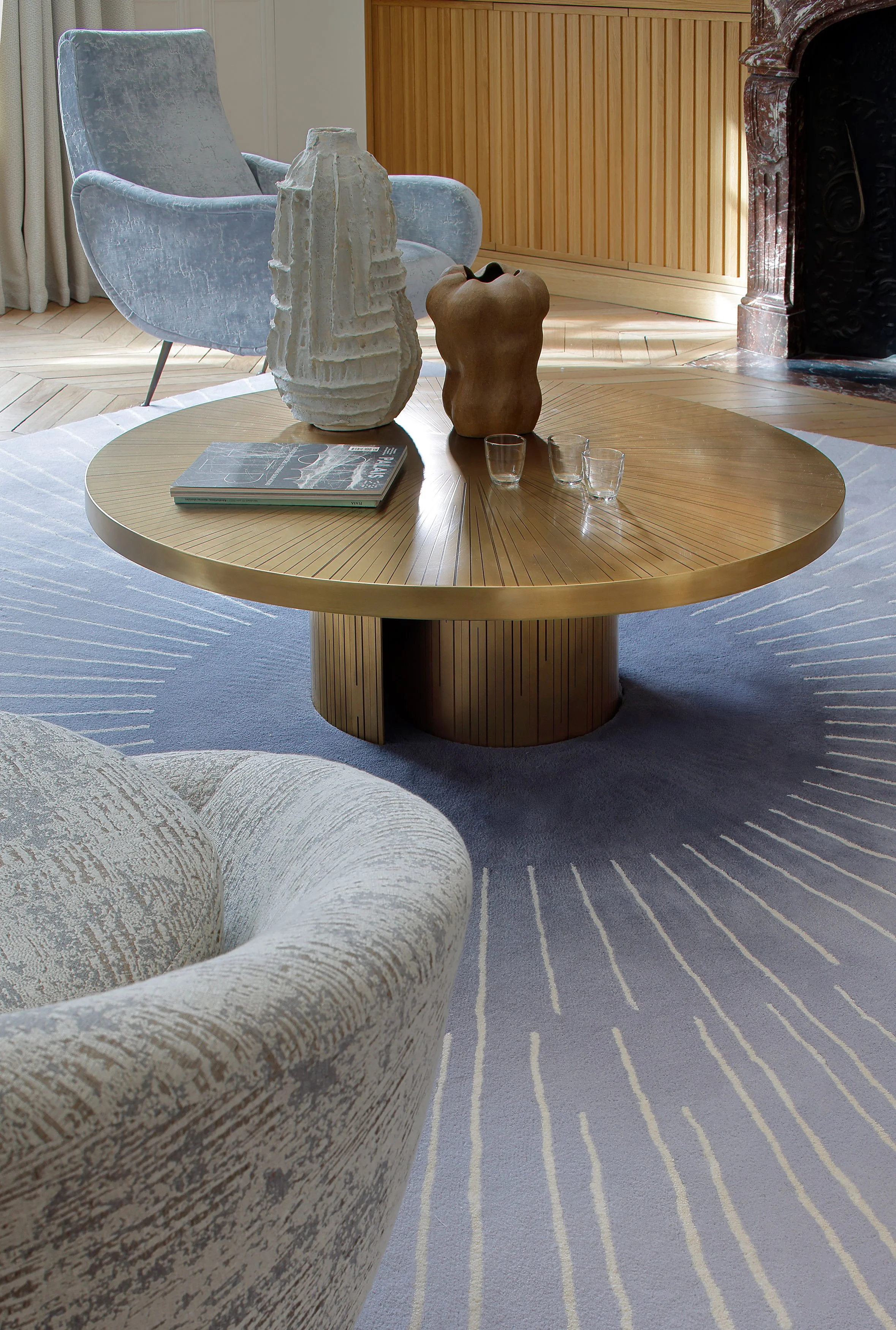 Table basse en bois doré avec deux sculptures modernes et trois verres, dans un salon élégant avec un fauteuil en velours gris clair, un tapis à rayures blanches et une cheminée en marbre.