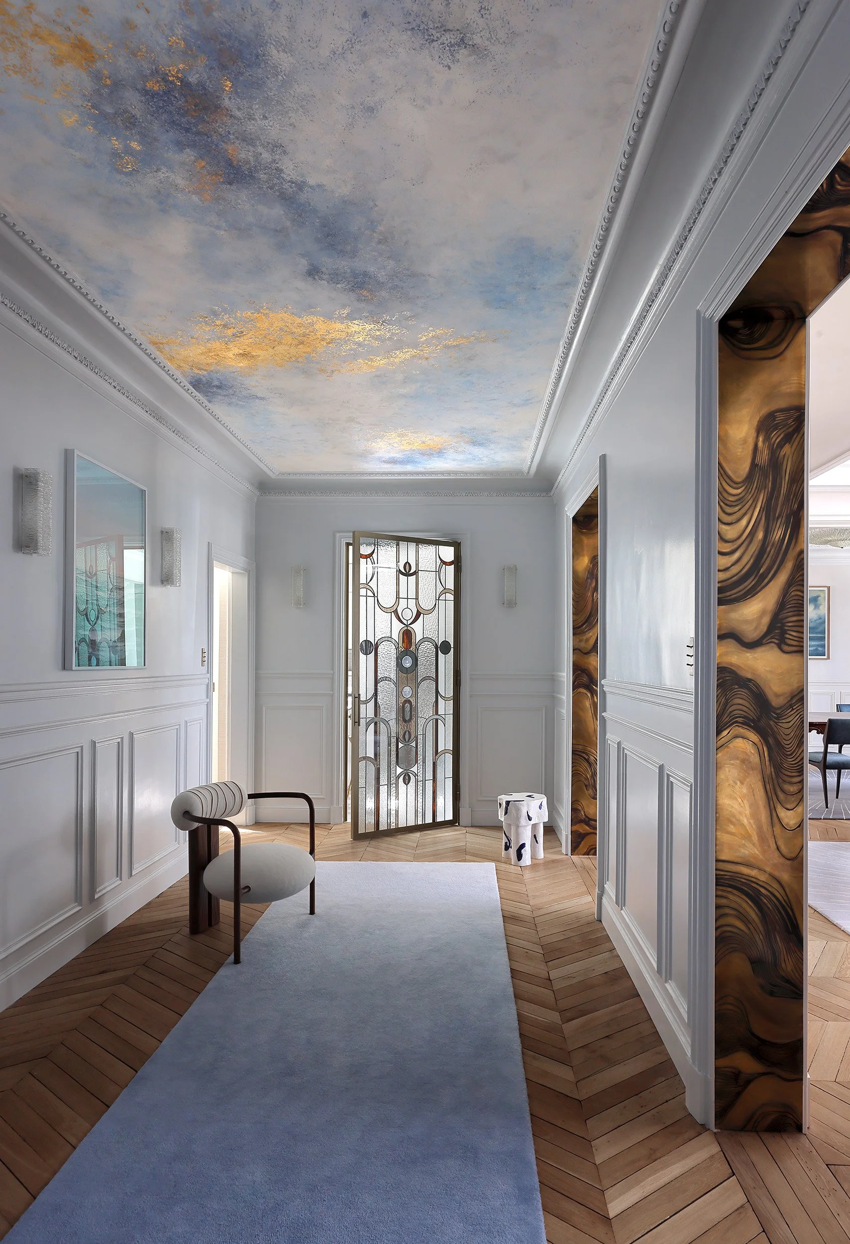 Entrée avec plafond peint en ciel bleu et nuages, sol en parquet, porte en verre décorative, un fauteuil blanc avec cadre en bois à gauche, mur blanc avec moulures, une table en forme de vache en blanc et noir, mural de marbre marron sur le côté droit.