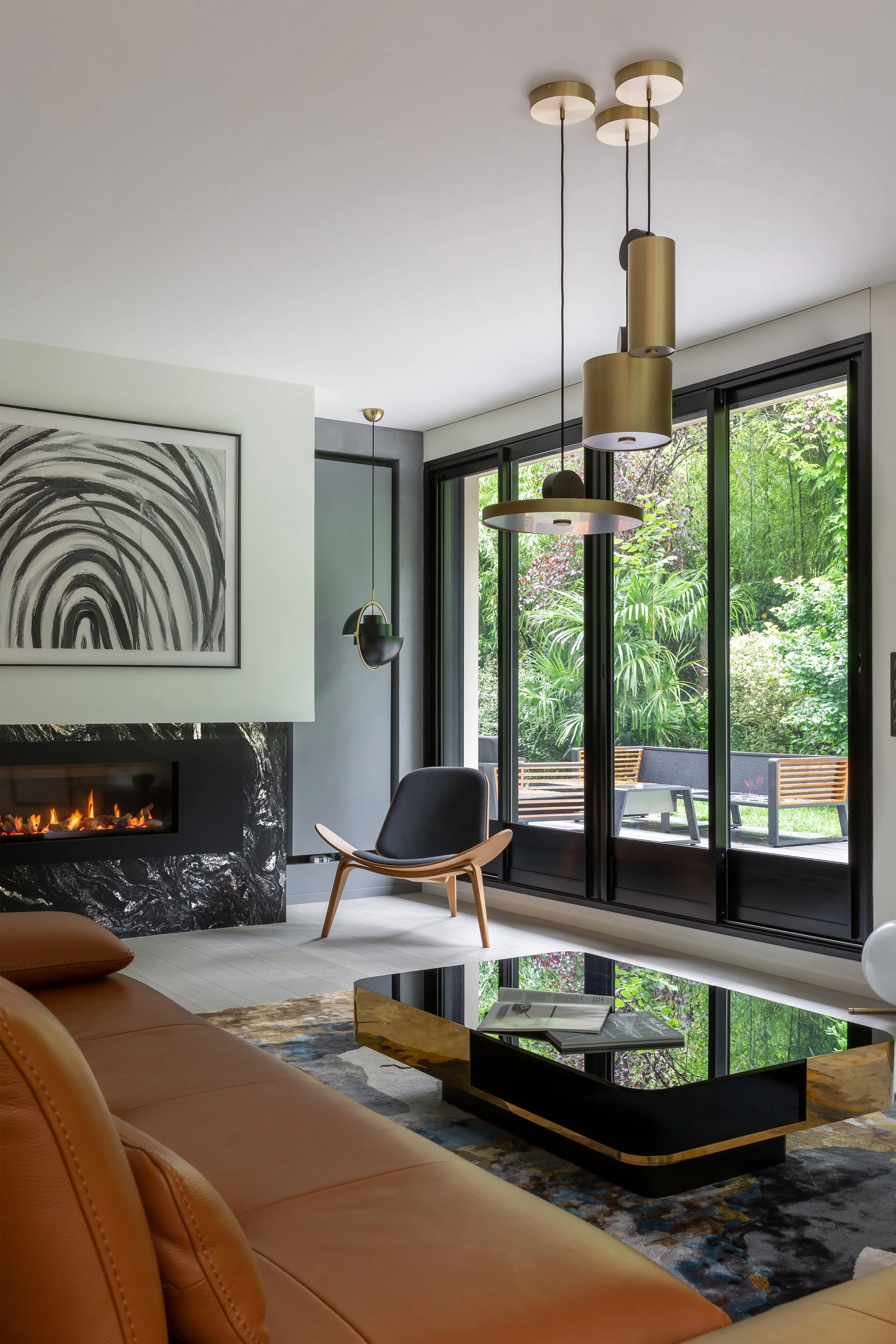 Salon moderne avec cheminée, fauteuil, table basse en noir et or, grande baie vitrée donnant sur un espace extérieur verdoyant.
