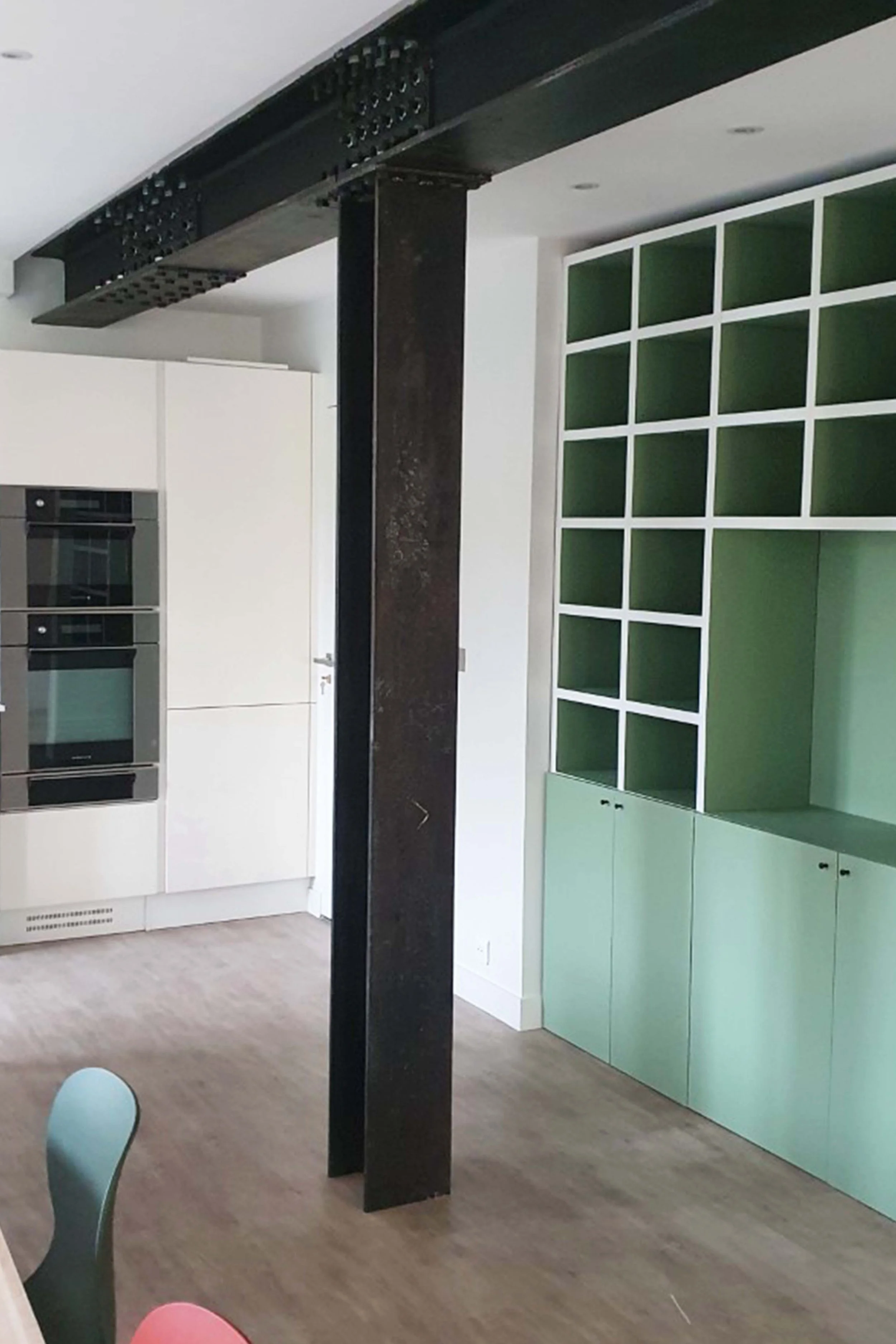 Interior of a modern kitchen with white cabinets, a wall with green shelves, and a black metal column.