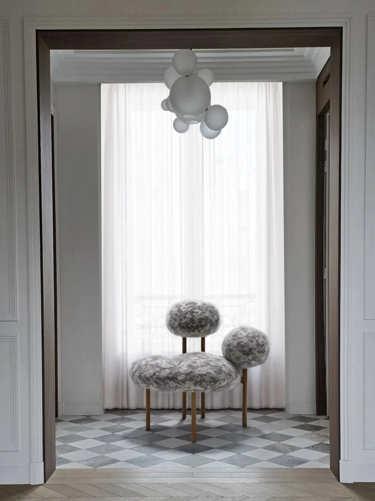 Une pièce lumineuse avec un fauteuil en peluche gris, un rideau blanc, un plafond suspendu avec un lampadaire en forme de sphères blanches, murs blancs et sol en carreaux gris et blanc.
