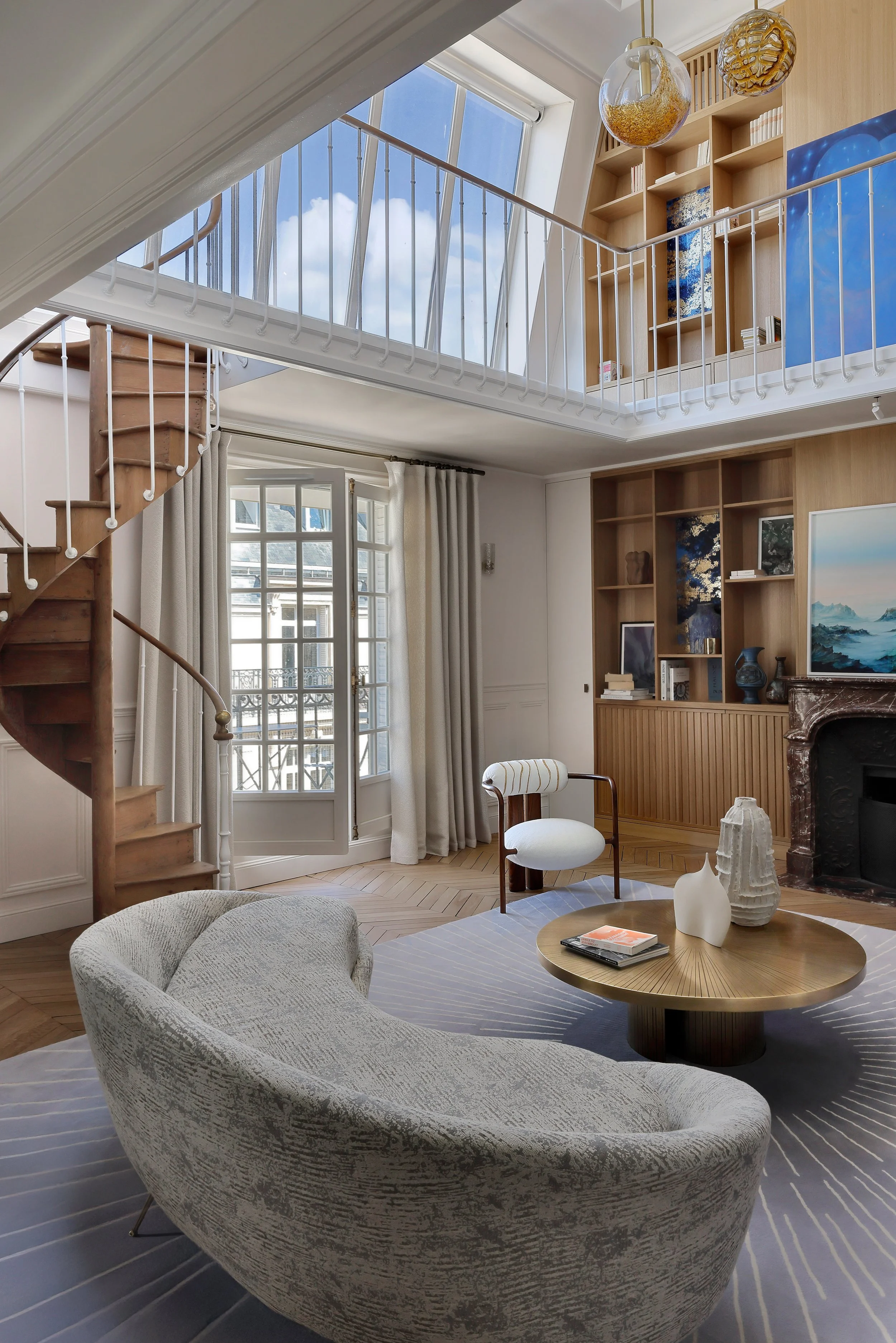 Salon lumineux avec canapé beige, chair blanche, table ronde en bois, cheminée en marbre, étagères en bois avec décorations, grande fenêtre avec rideaux, escalier en bois, et plafond avec de grands fenêtres et deux luminaires