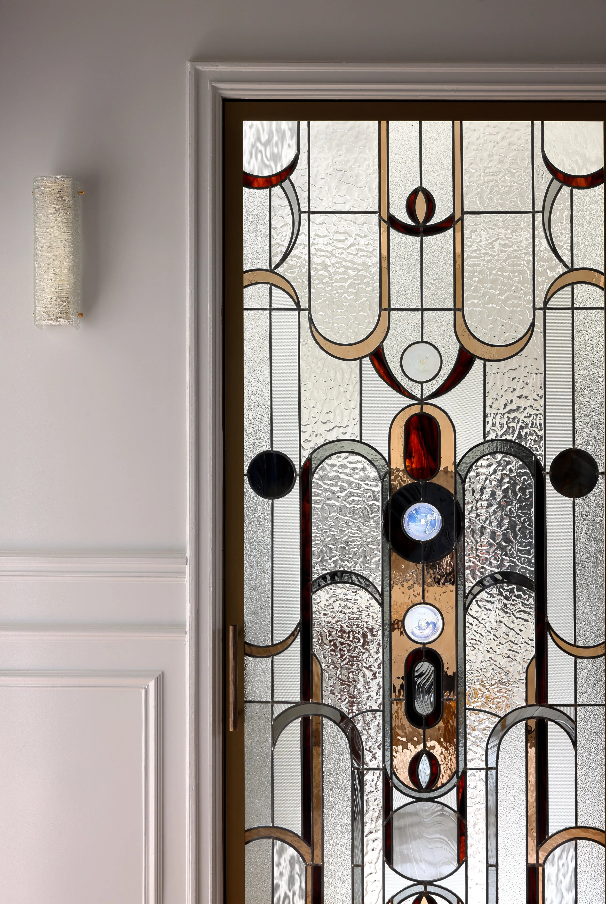 Une porte en verre avec un motif design en métaux et verre coloré, encadrée par une moulure blanche dans un mur blanc, avec un luminaire mural placé à gauche.