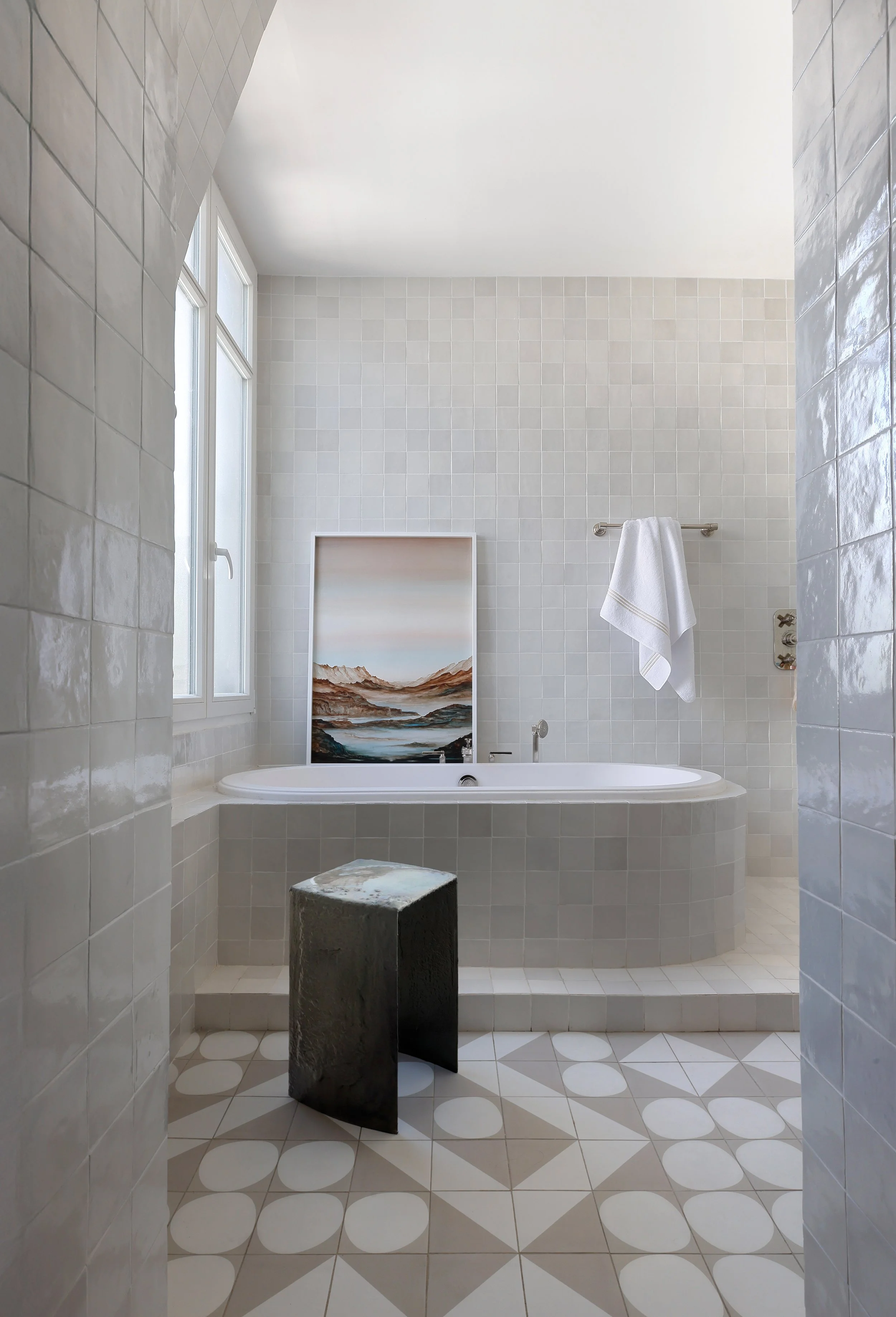 Salle de bain avec une baignoire, tableau, serviette blanche, et sol en carrelage géométrique beige et blanche.