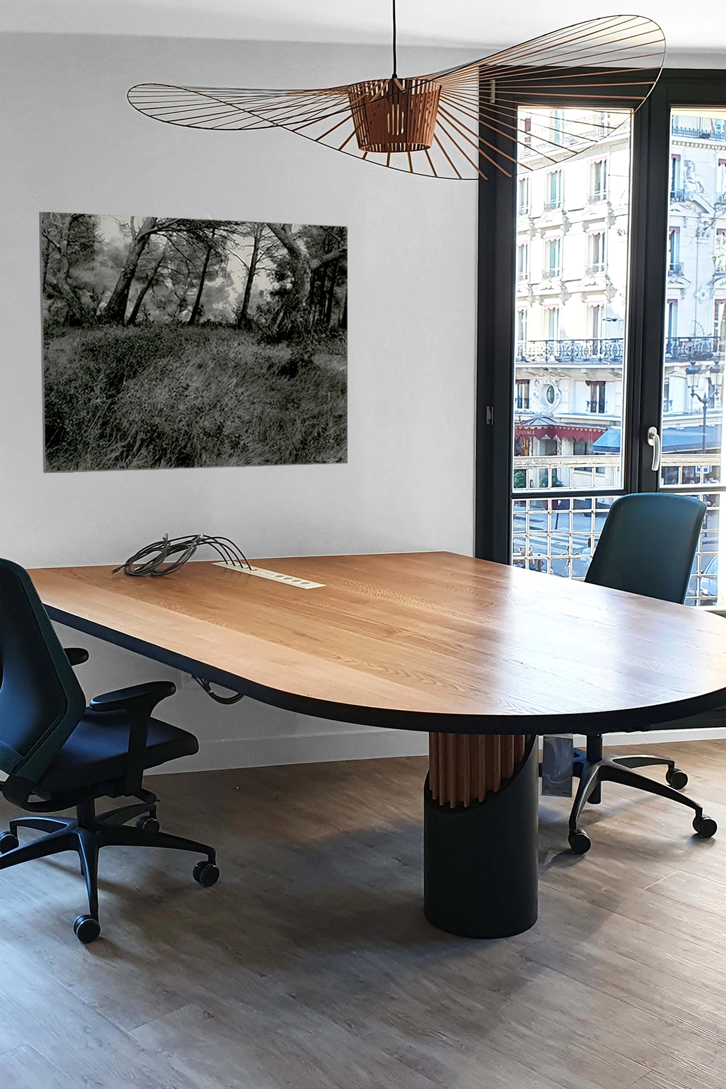 Une salle de réunion avec une grande table en bois, deux chaises de bureau, un grand panneau d'affichage avec une photo en noir et blanc d'une forêt, et une grande fenêtre laissant passer la lumière naturelle.