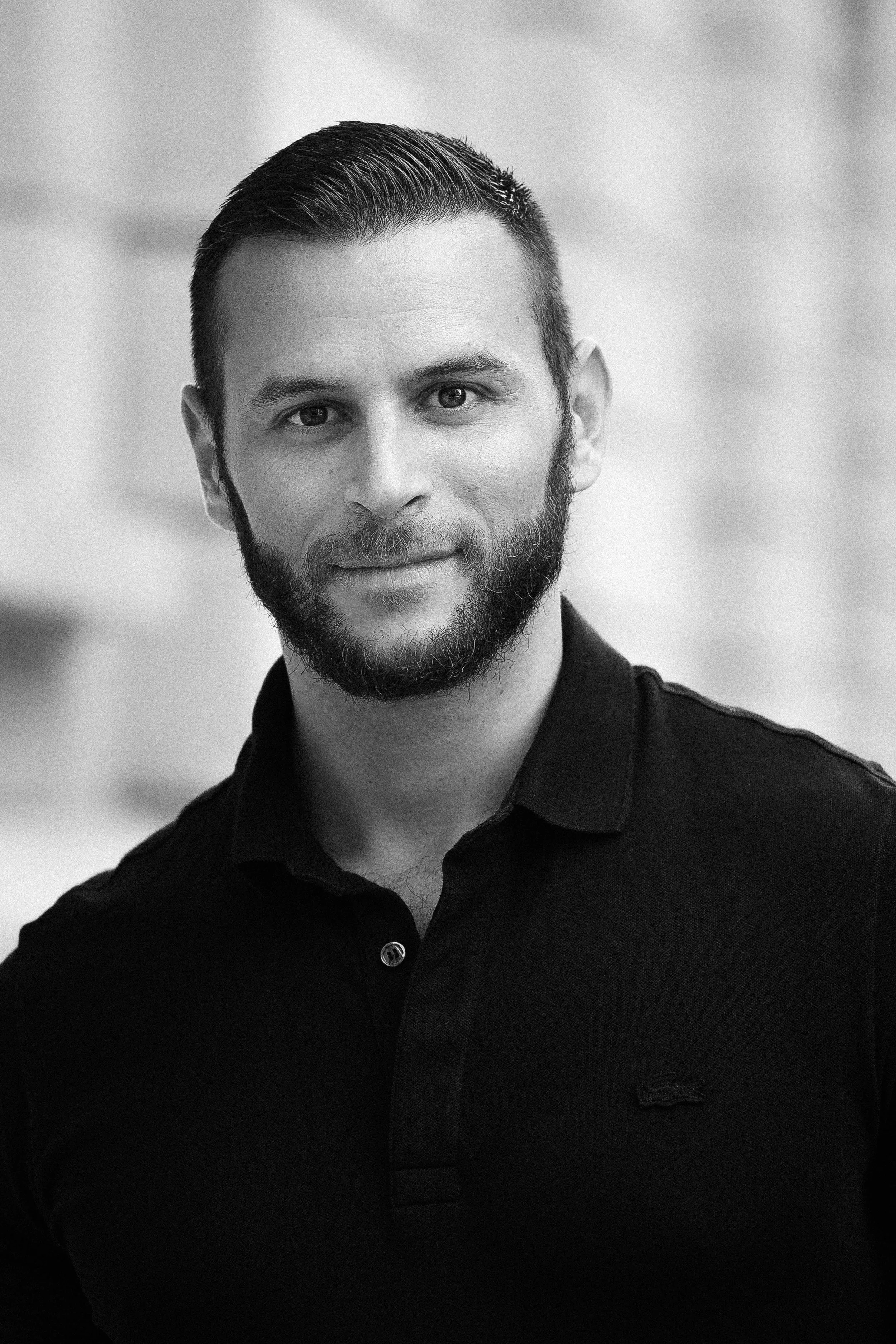 Portrait en noir et blanc d'un homme (Sébastien Coudert-Maugendre) avec une barbe, portant une chemise noire, regardant l'objectif avec un léger sourire.