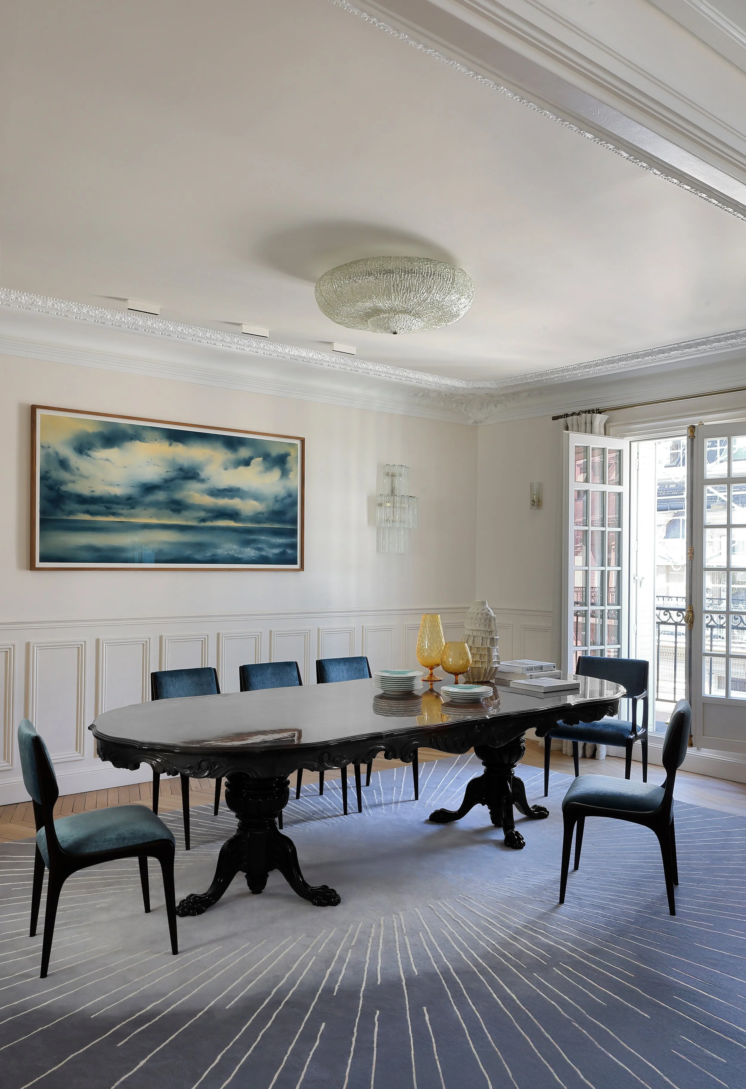Une salle à manger élégante avec une table en bois sombre, six chaises en tissu bleu foncé, un grand tableau représentant un ciel orageux, un plafond avec un luminaire en cristal, une fenêtre avec une porte-fenêtre donnant sur un balcon, et une décoration avec des vases en verre et des livres.