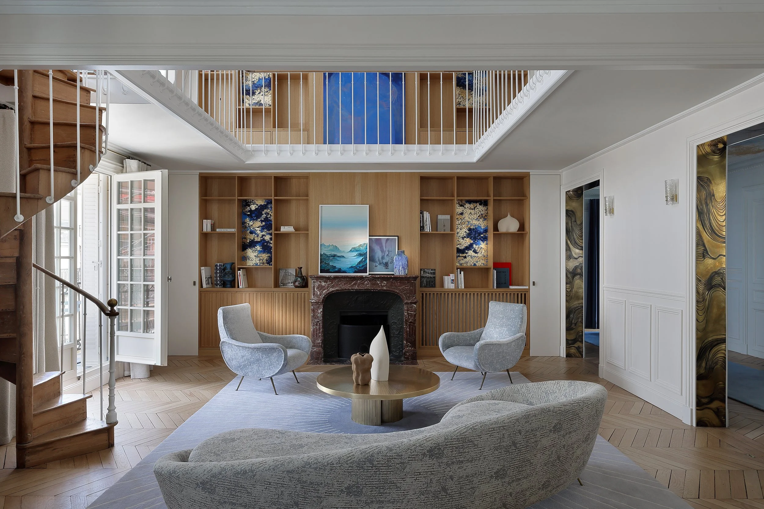 Salon avec cheminée en marbre, deux fauteuils gris, canapé gris, table basse en bois avec vases, bibliothèque en bois avec décoration