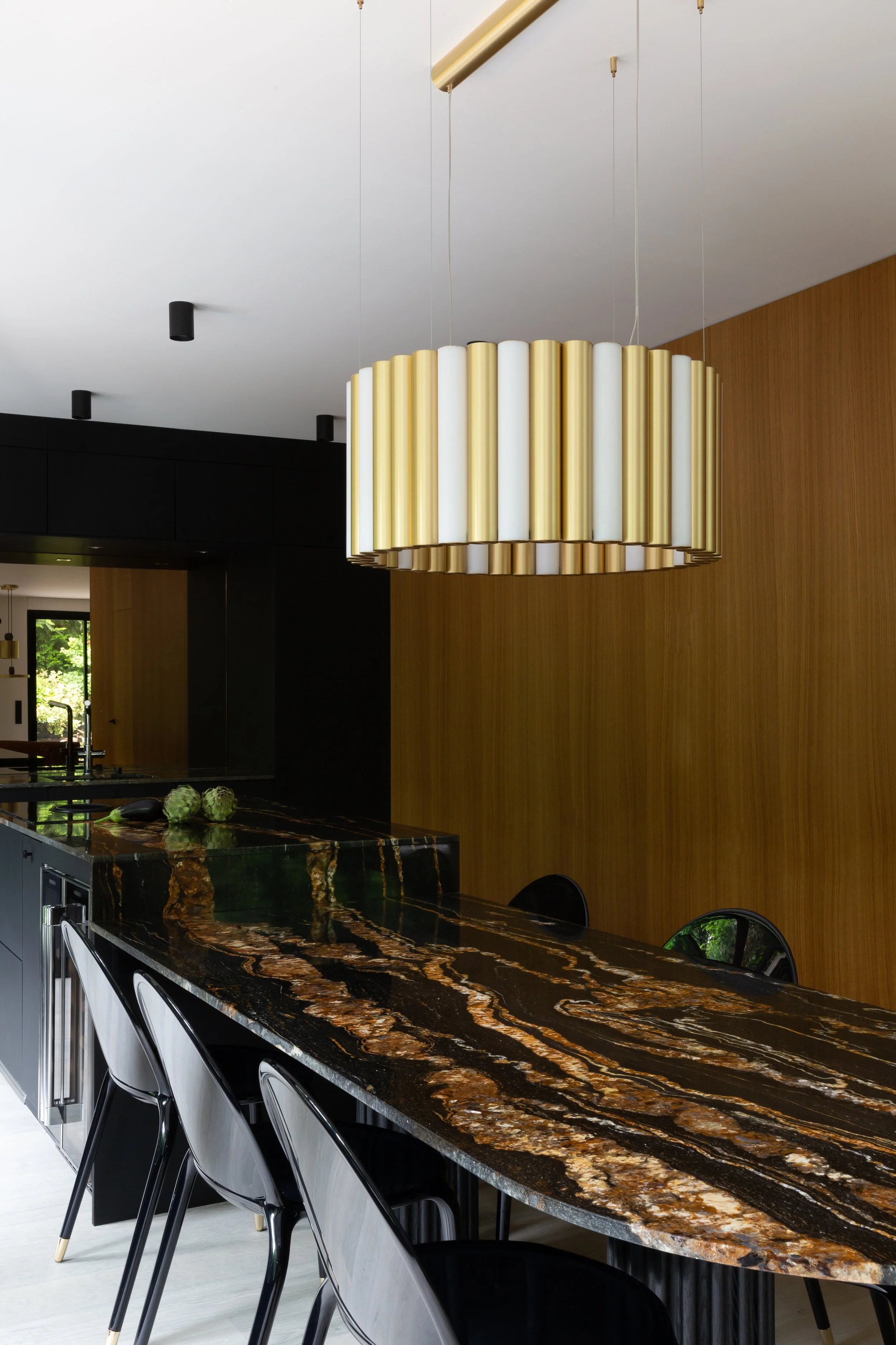 Un panneau de cuisine avec un plan de travail en pierre noire et marron, un grand lustre suspendu en métal doré et blanc, et des chaises noires