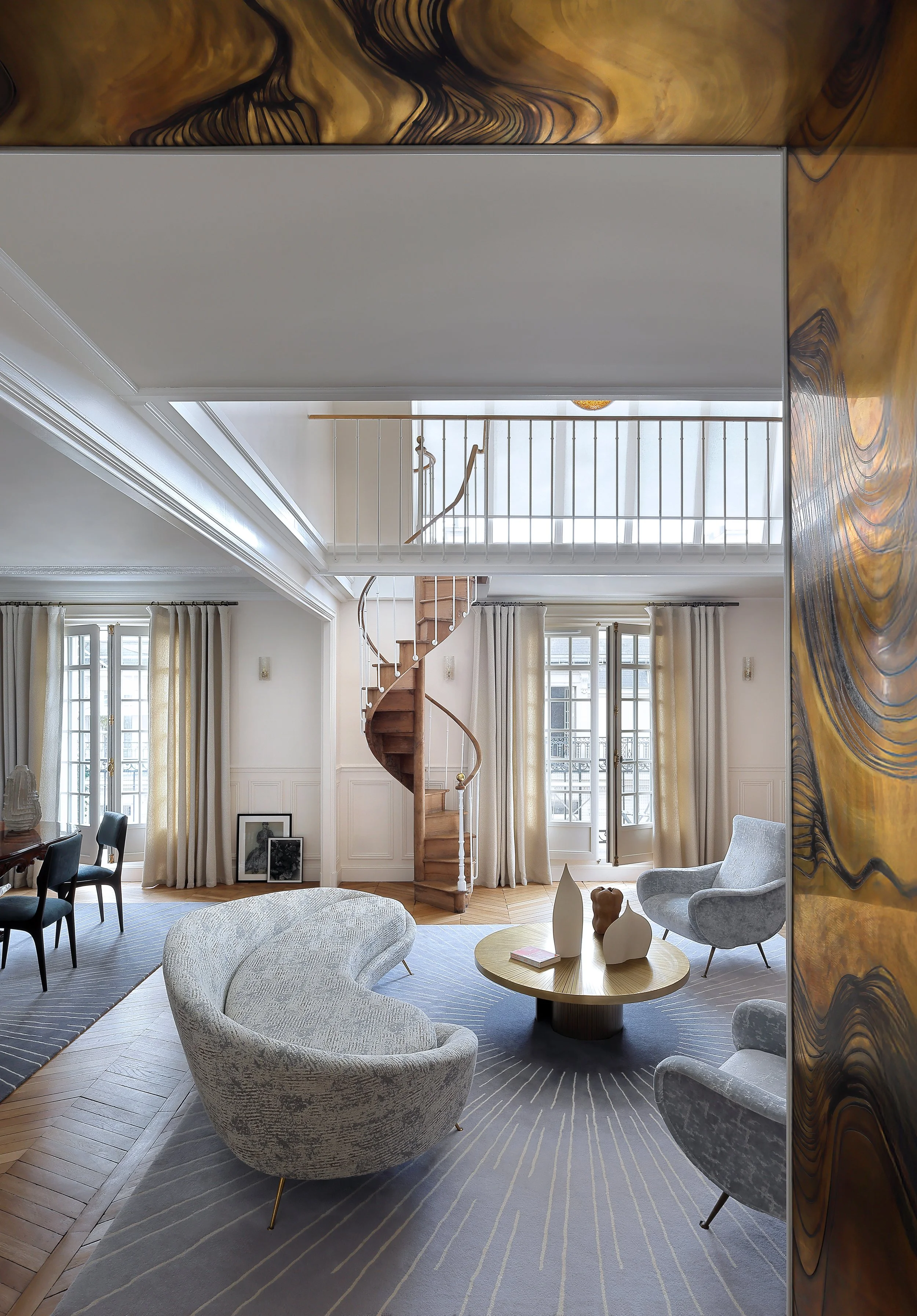 Salon moderne avec canapé en forme de demi-cercle, fauteuils en tissu gris, tapis rayé, grande table basse ronde en bois, murs blancs, fenêtres avec rideaux écrus, escalier en spirale en bois, balcon intérieur avec rampe blanche, décorations en céramique sur la table.