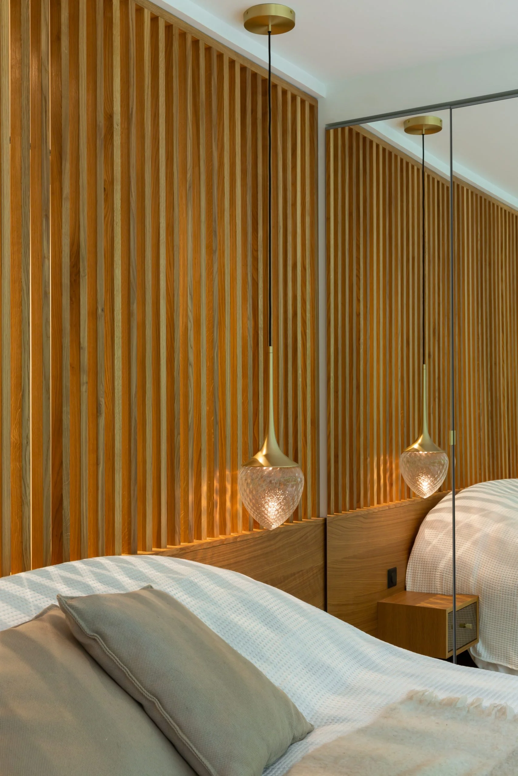 Bedroom with striped wood wall and two hanging lamps in glass and gilded metal, mirror in mirror-holder and bed with white sheet and beige pillows.