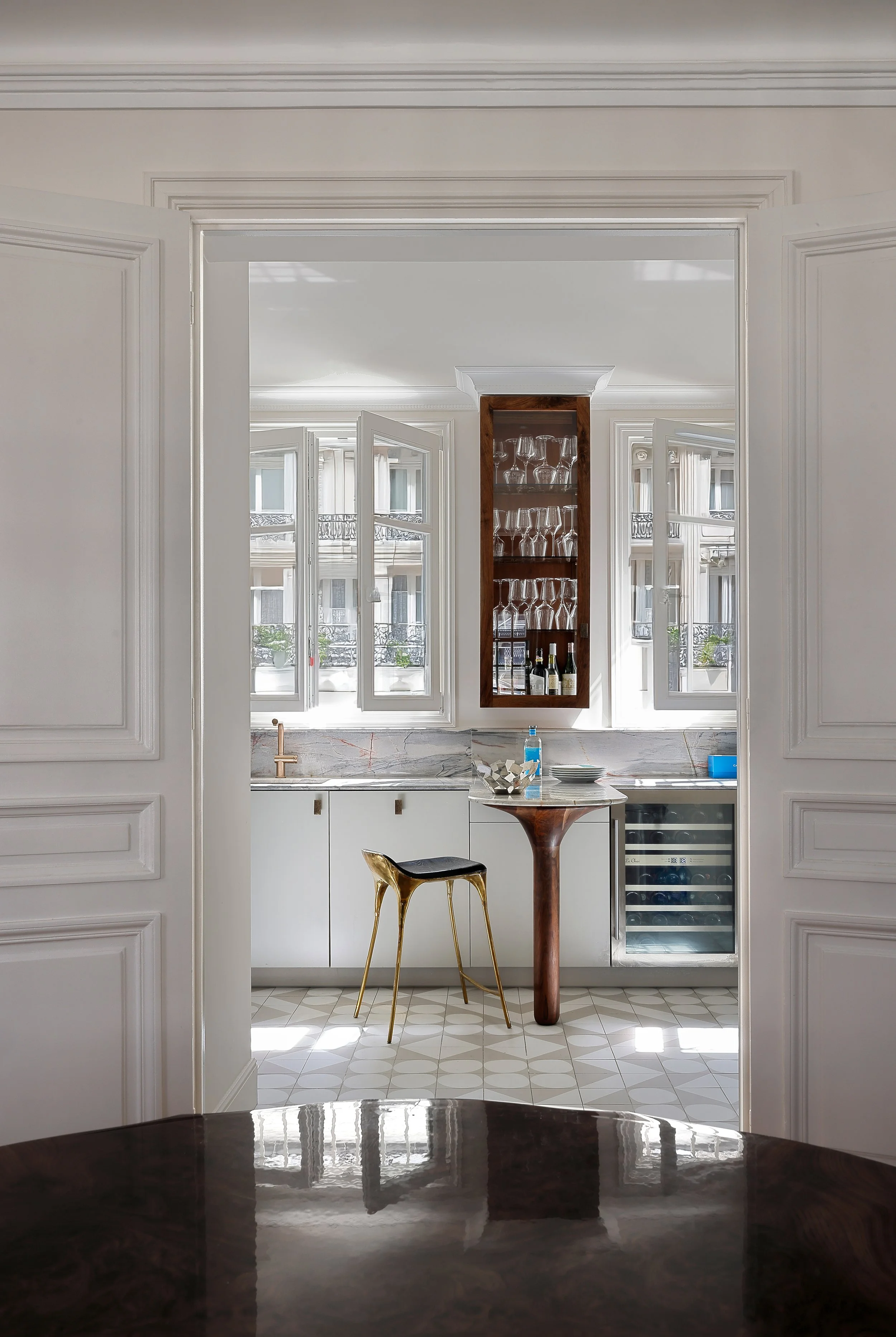Une vue d'une cuisine lumineuse avec des fenêtres ouvertes, un meuble en bois avec des verres à vin, une bouteille de vin et un distributeur de savon bleu, une table en bois avec une chaise dorée, et un sol à motifs géométriques.