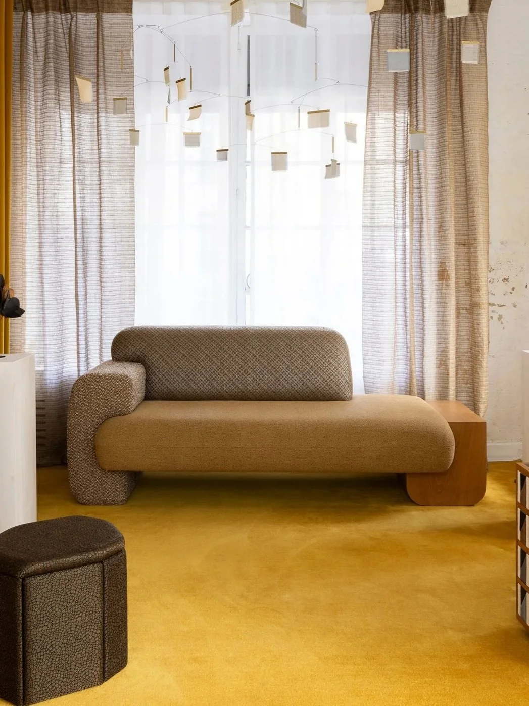 Un salon avec un canapé beige et un fauteuil noir sur un tapis jaune. Il y a des rideaux à rayures derrière une fenêtre avec des voilages blancs, et un mobile suspendu en haut au centre de l'image.