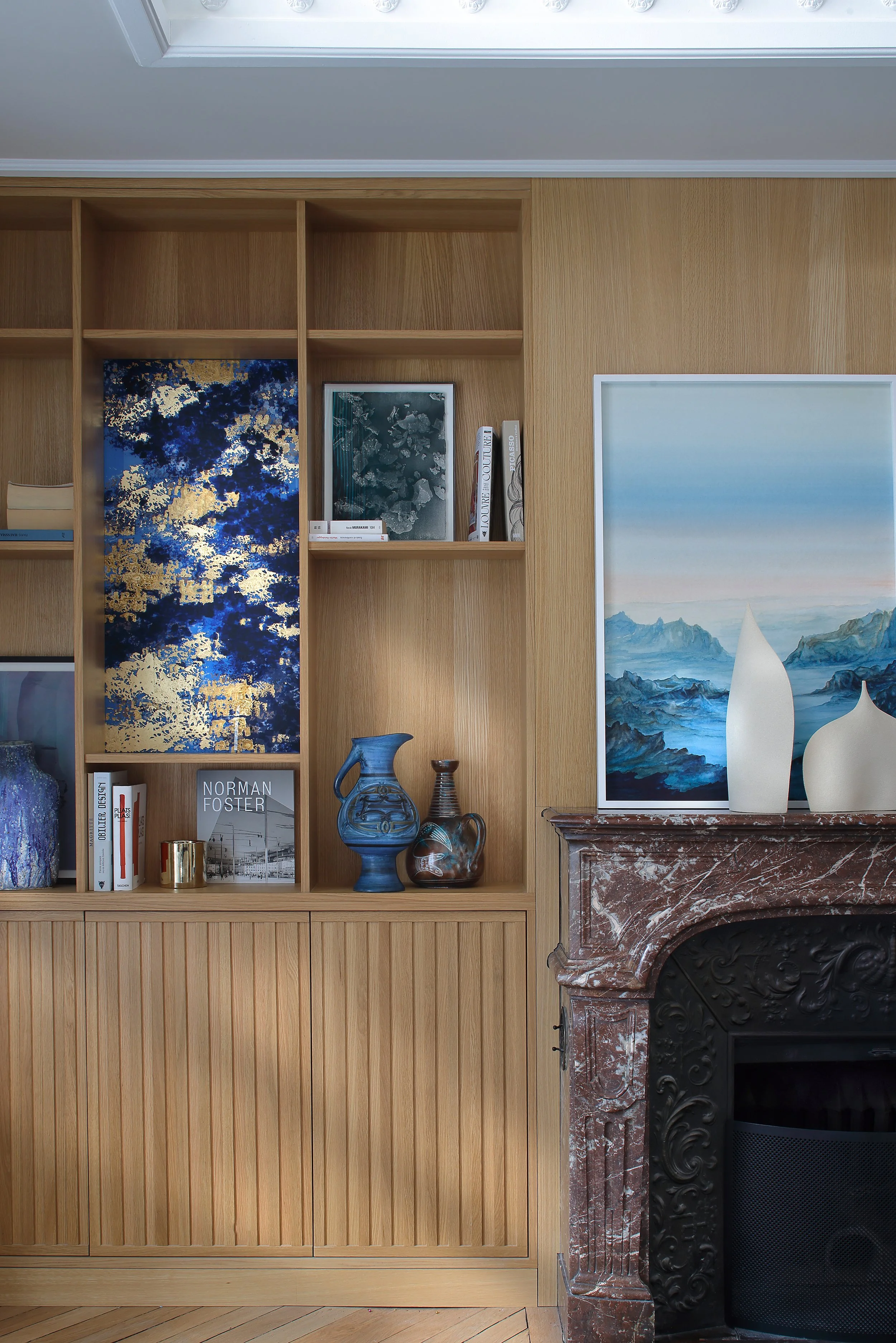 Une cheminée en marbre rougeâtre avec des vases blancs sur le dessus, un mur en bois avec étagères contenant des livres et des objets décoratifs, et une peinture représentant un paysage côtier attachée au mur.